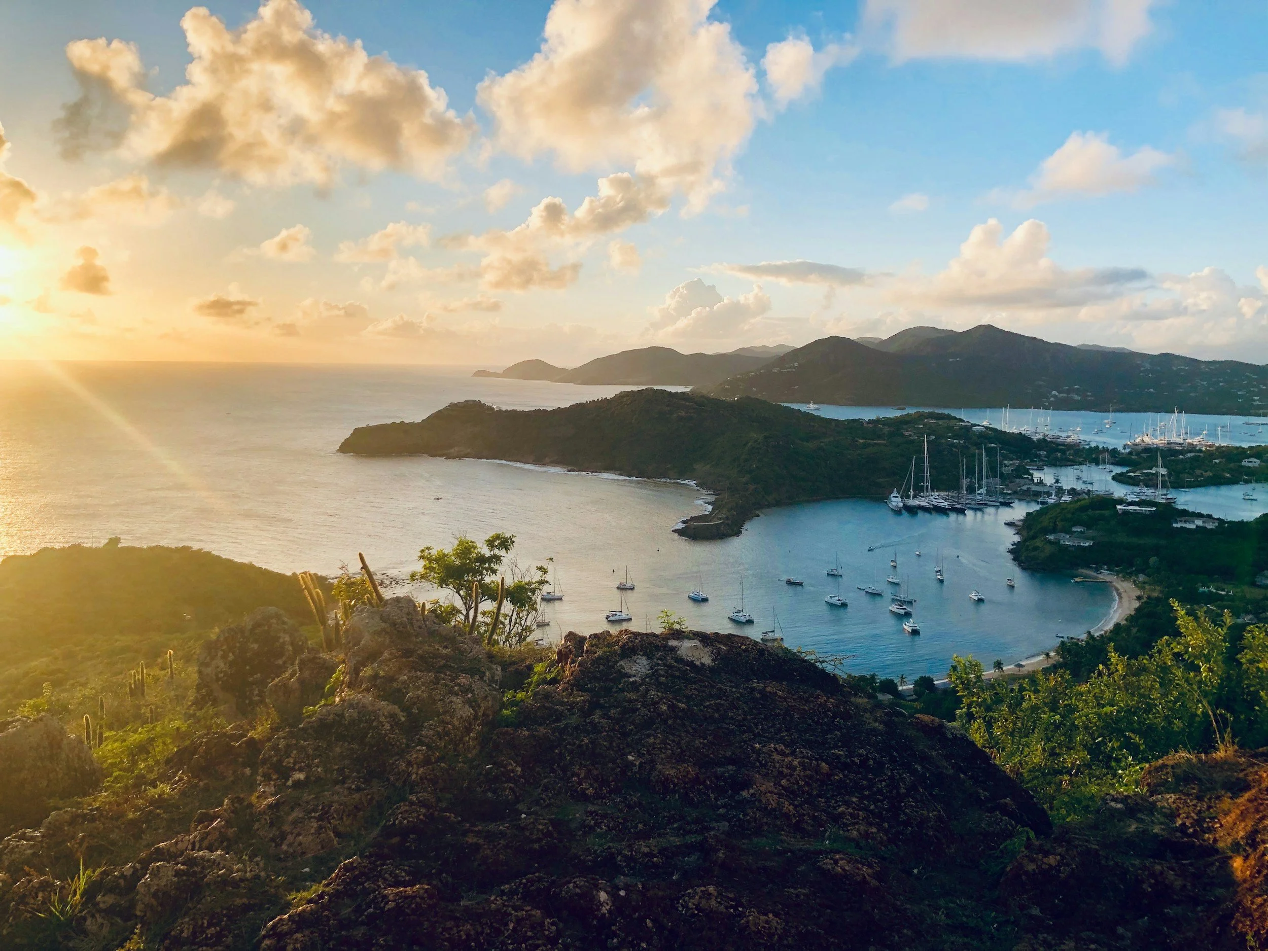 Scenic view of English Harbour from a hilltop, featuring a blue ocean, anchored sailboats, and lush green hills at sunset.