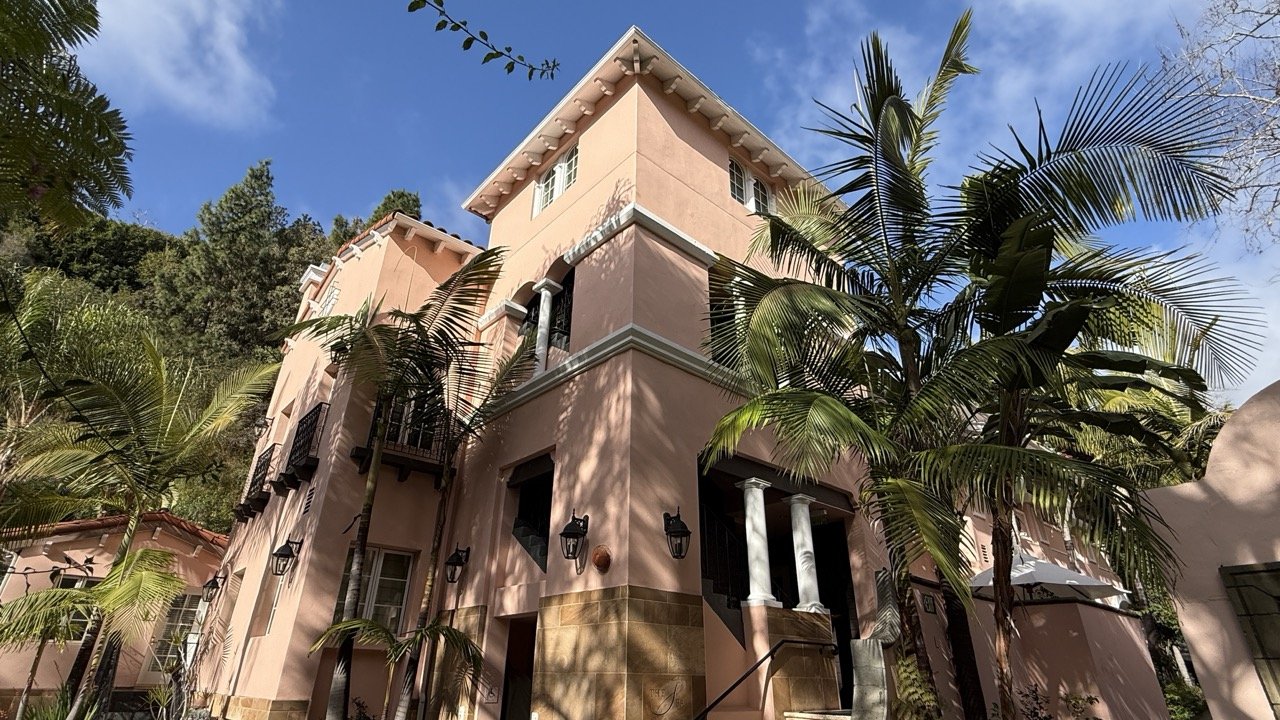 Exterior view of Hotel Bel-Air in Los Angeles, featuring pastel pink architecture, lush palm trees, and a clear blue sky – captured by The Private Traveller.