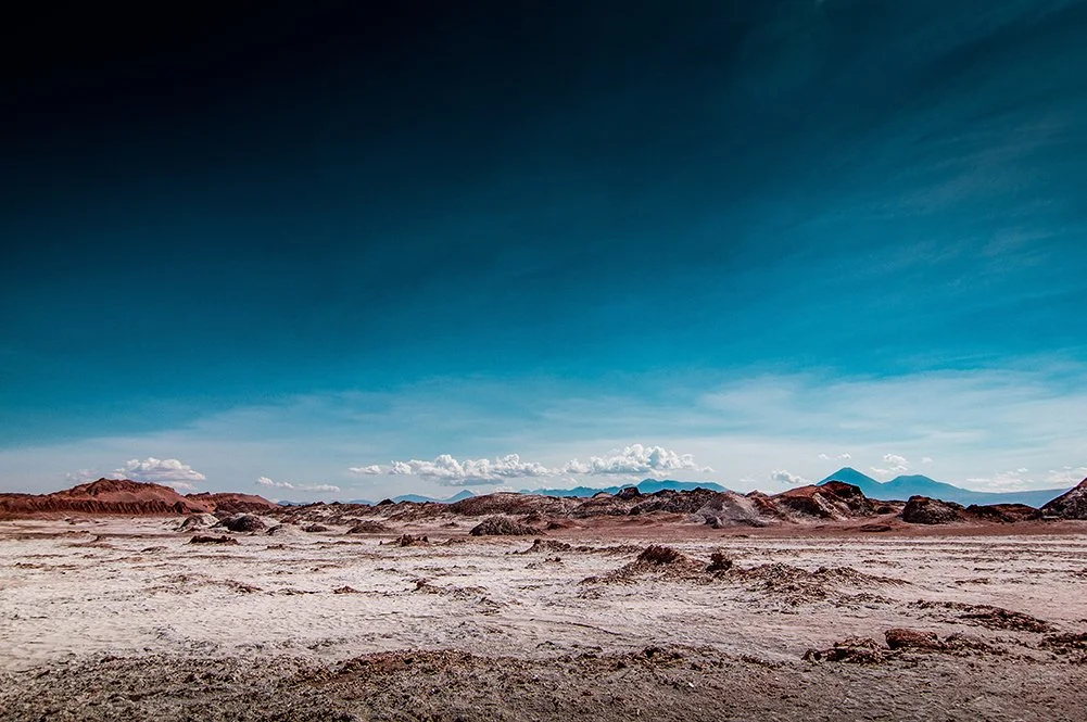 Desert landscape with dusty ground and distant mountains under a bright blue sky.