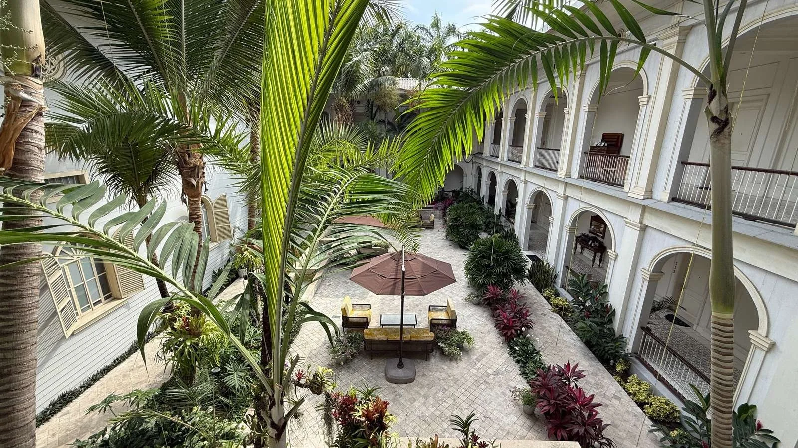 Upper-level balcony view across a serene tropical courtyard with palms and colonial arches
