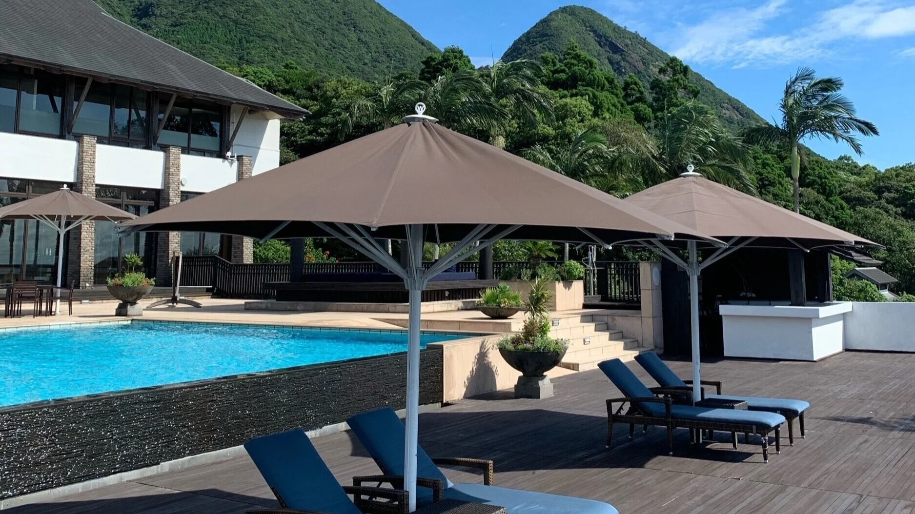 Bright blue swimming pool with wooden deck and loungers in front, hotel building behind in front of luscious green mountains