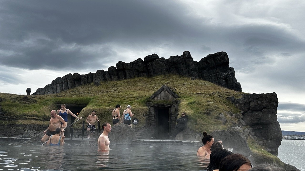 Steam coming off thermal pool with people walking around with grass and rocky backdrop