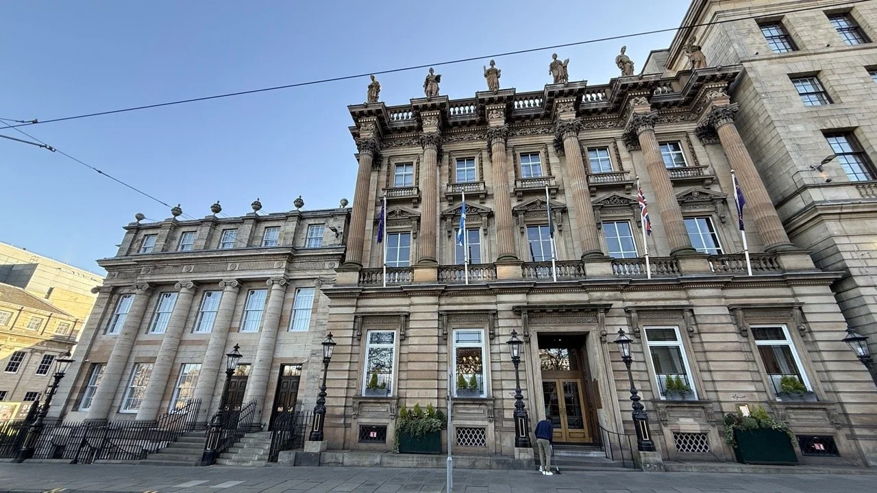 The grand neoclassical facade of Gleneagles Townhouse in Edinburgh, featuring towering columns, intricate stonework, and national flags against a clear blue sky.