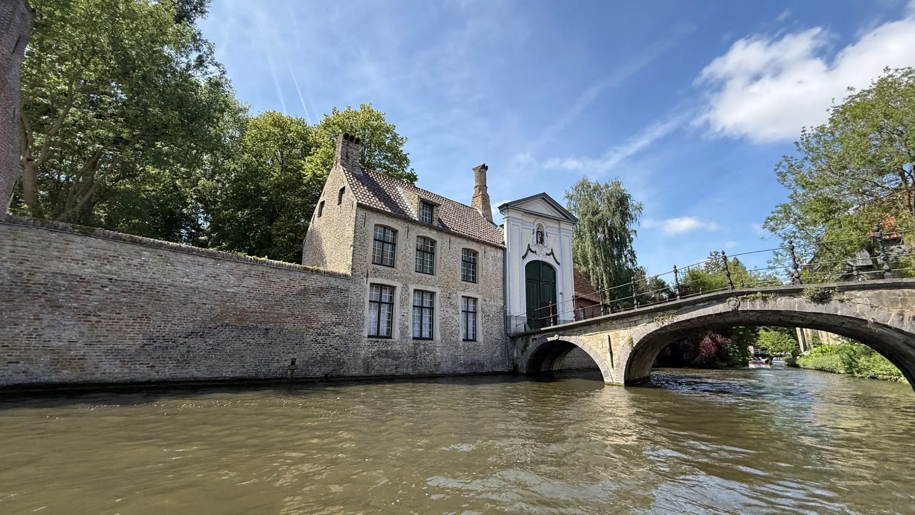 Historic canal bridge and traditional brick house beside the waterways of Bruges, Belgium, photographed during a canal boat tour on a sunny summer day.