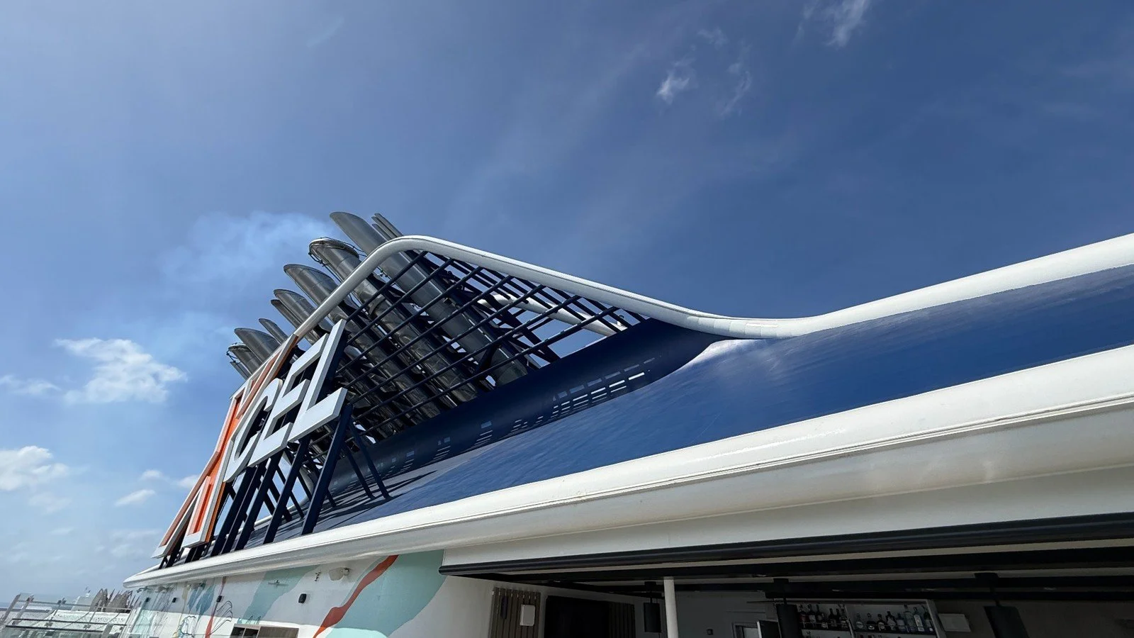Celebrity Xcel cruise ship funnel and upper deck photographed from below, showcasing the modern curved design, bold ‘Xcel’ branding and blue sky background.