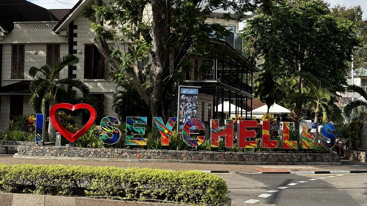 A vibrant “I ❤️ Seychelles” sign in Victoria, Seychelles, featuring colourful letters and a red heart, set against a backdrop of tropical plants and colonial-style buildings.