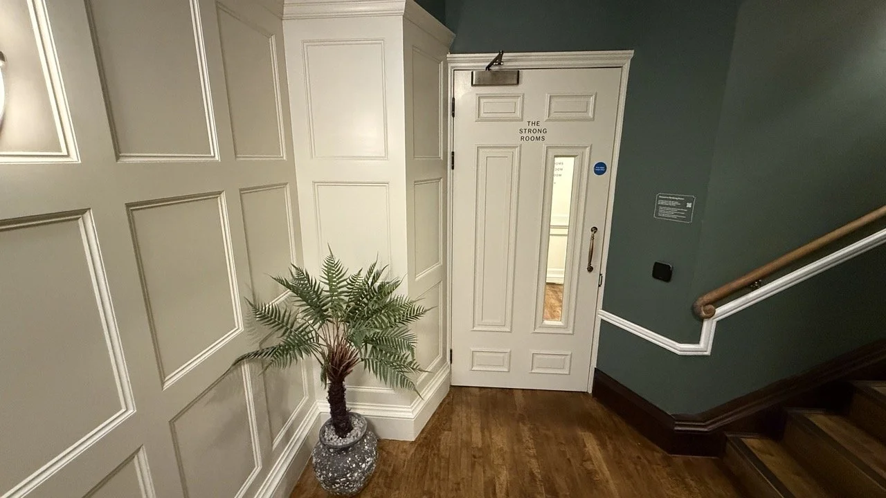 Elegant white entrance door with 'The Strong Rooms' signage, framed by dark green panelled walls and a potted plant.