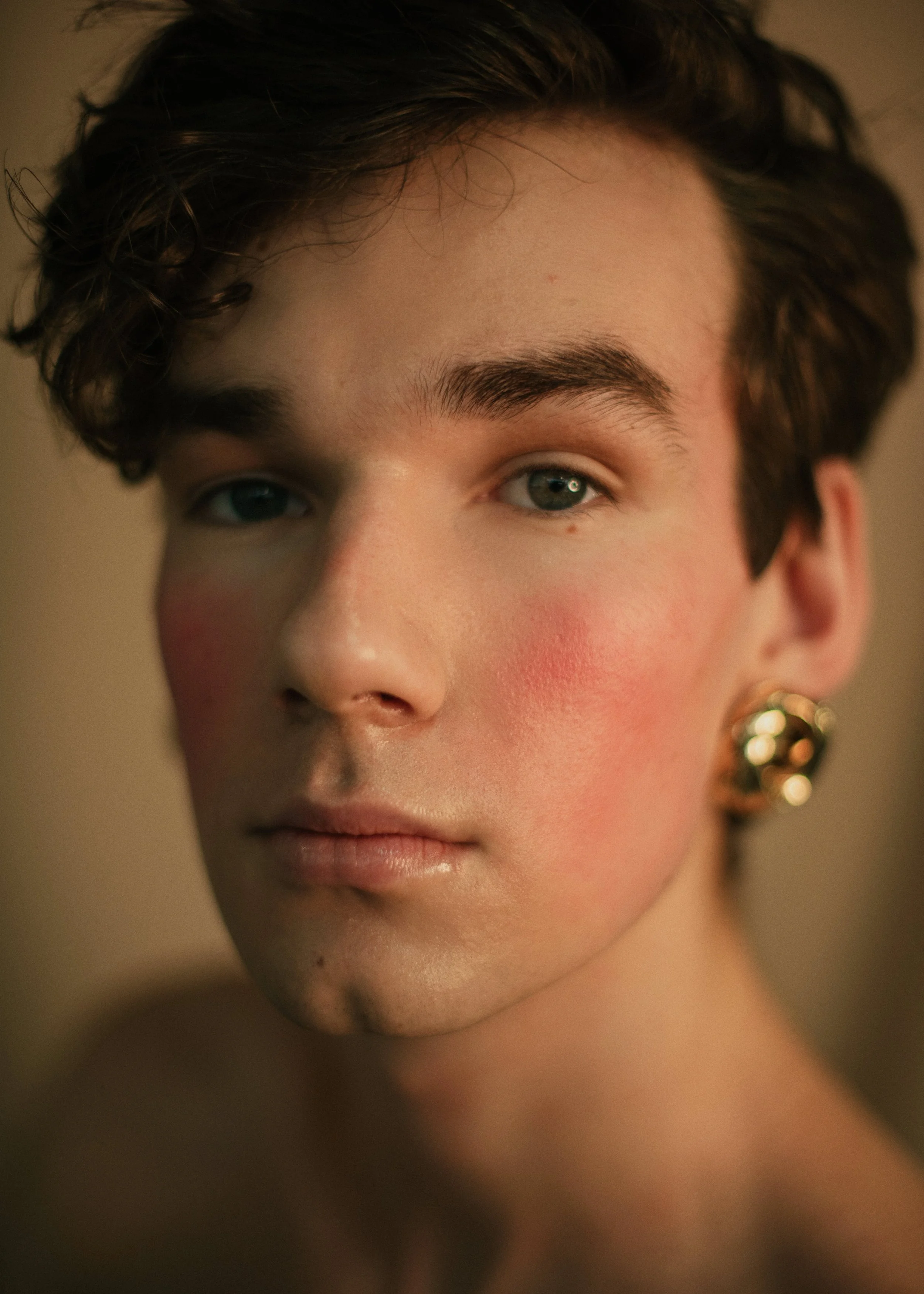 Portrait d'une personne queer aux cheveux bouclés, portant des boucles d'oreille dorées, sans vêtement et qui vous regarde