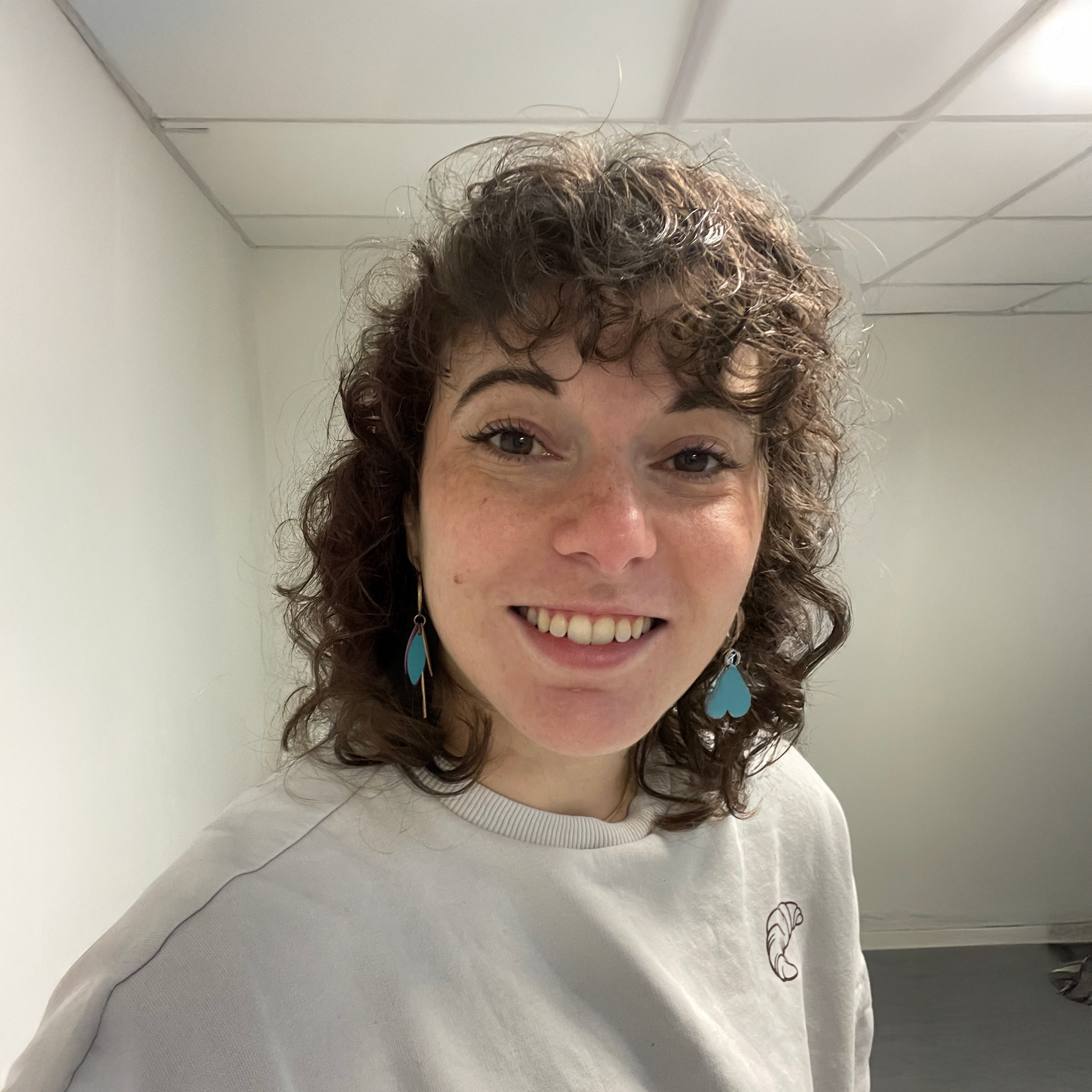 A woman with curly brown hair smiling at the camera, wearing turquoise heart-shaped earrings and a white sweatshirt.