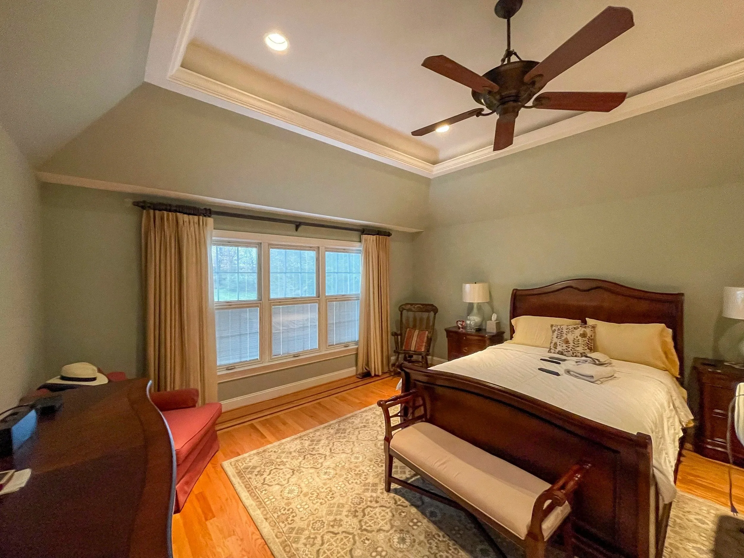A bedroom with a large wooden bed, beige curtains, and a sitting area with a red chair. The room has hardwood floors, a light green wall, and a tray ceiling with recessed lighting and a ceiling fan.