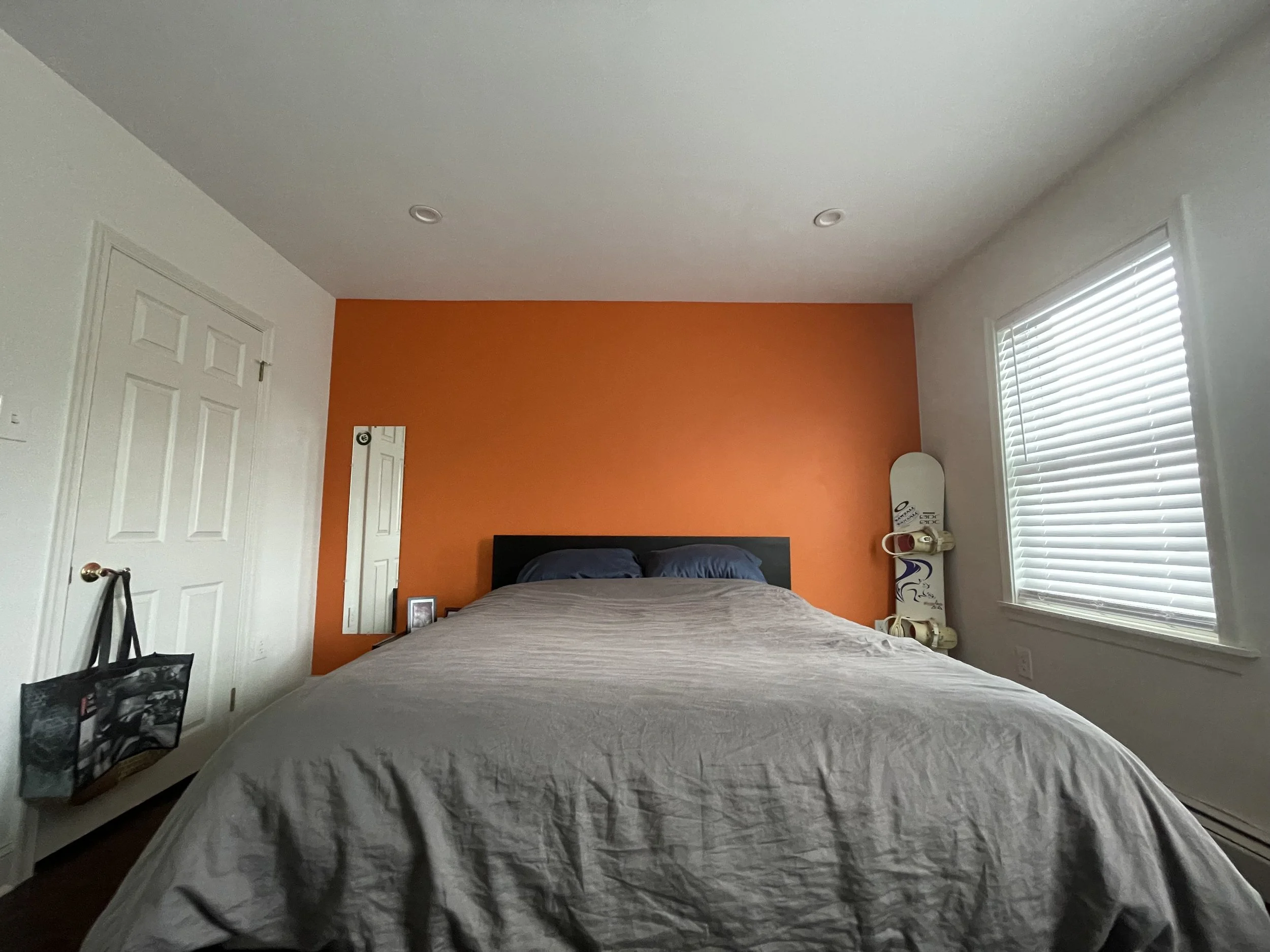 A bedroom with a large bed against an orange accent wall, two pillows, a full-length mirror, a small framed photo on a nightstand, a skateboard with shoes on it by the window, and a black tote bag hanging on a door handle.
