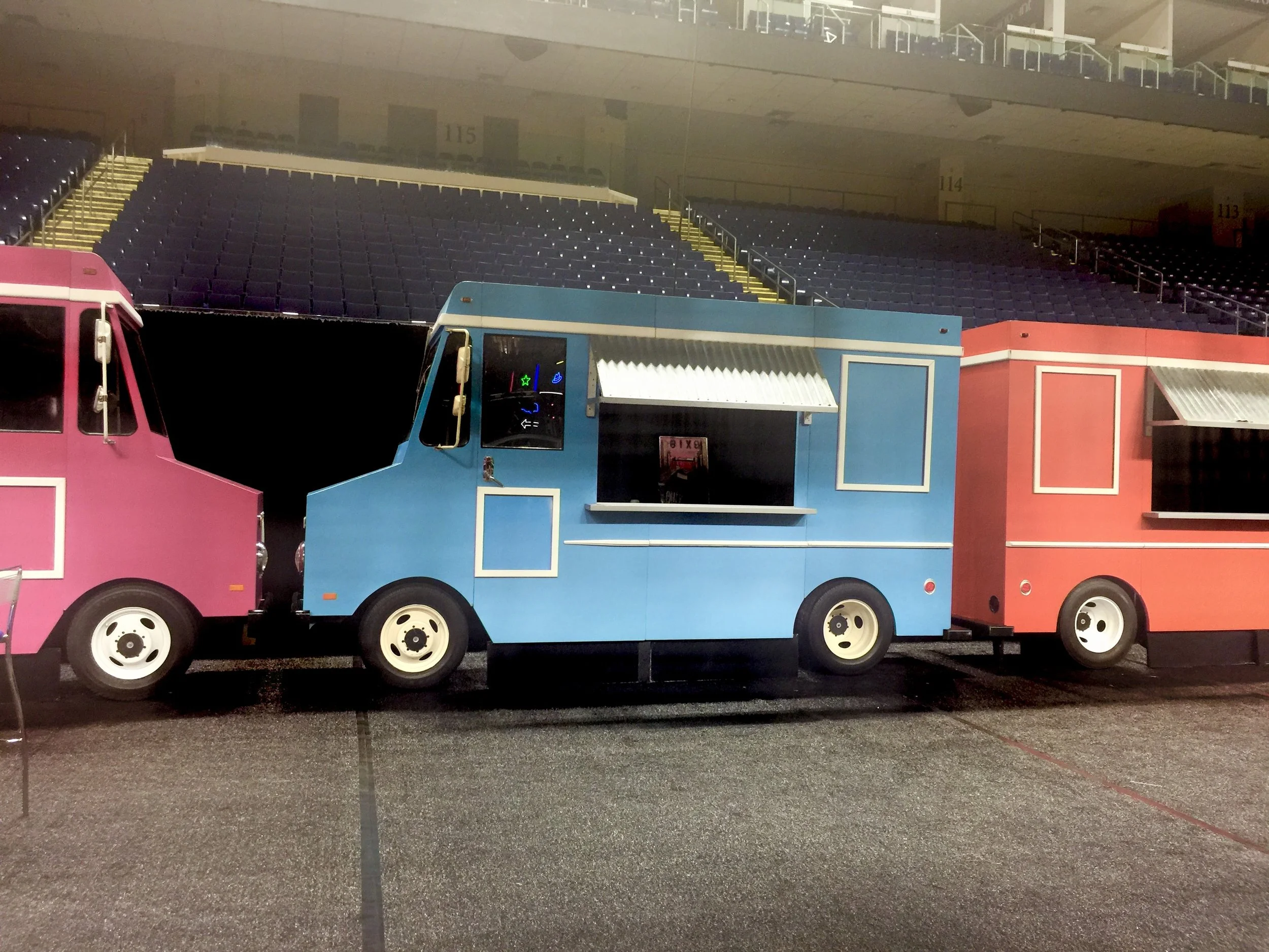 Three colorful food trucks, pink, blue, and red, parked inside a stadium with empty purple seats visible in the background.