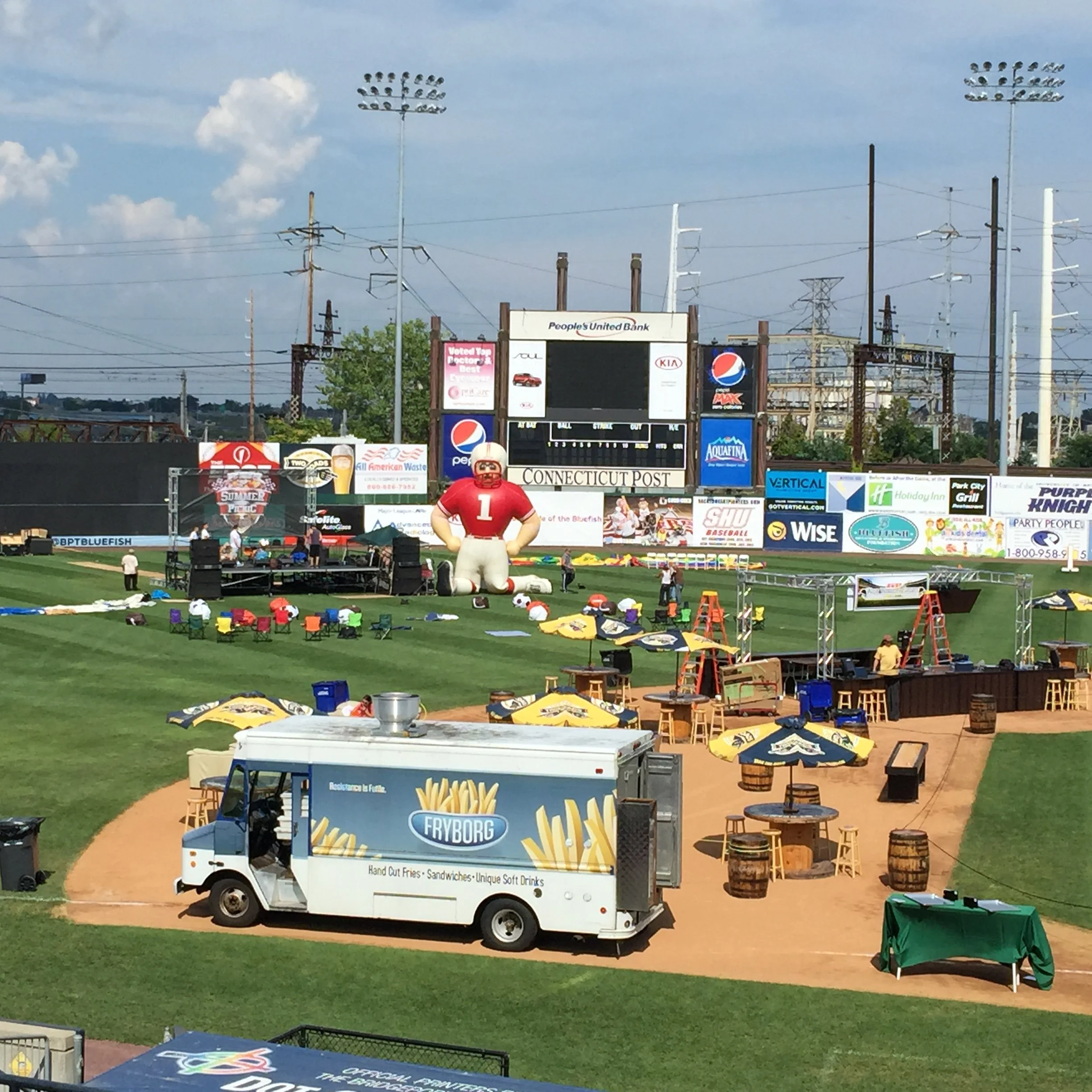 A baseball stadium under preparation with an inflatable football player figure, a Fryborg food truck, and various tables and umbrellas on the field, amidst equipment and scaffolding.