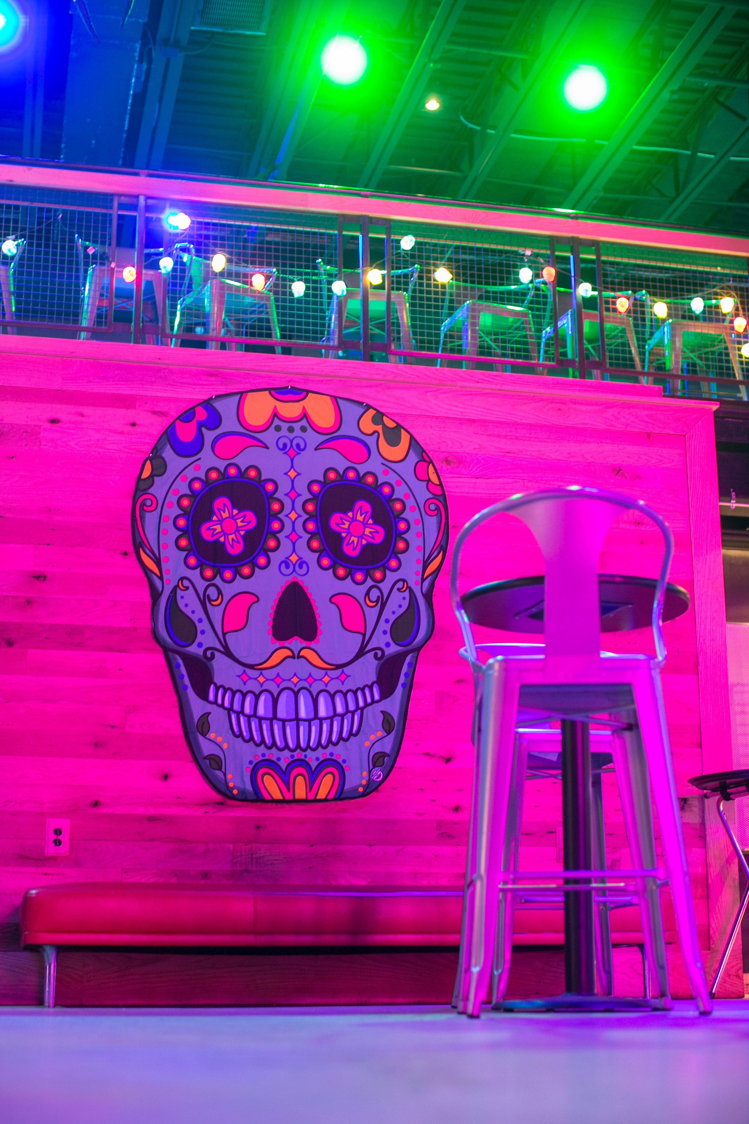 Decorated skull artwork on a pink wooden wall with string lights and high chairs in an indoor space with vibrant green lights overhead.