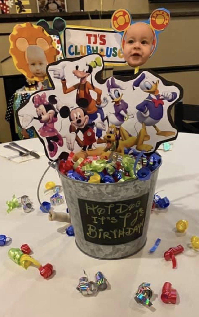 A birthday table centerpiece featuring a small metal bucket with a sign that says "Happy Dog IPtized Birthday!" Inside the bucket are colorful party decorations and cutouts of Disney characters Mickey Mouse, Minnie Mouse, Donald Duck, Daisy Duck, Goo