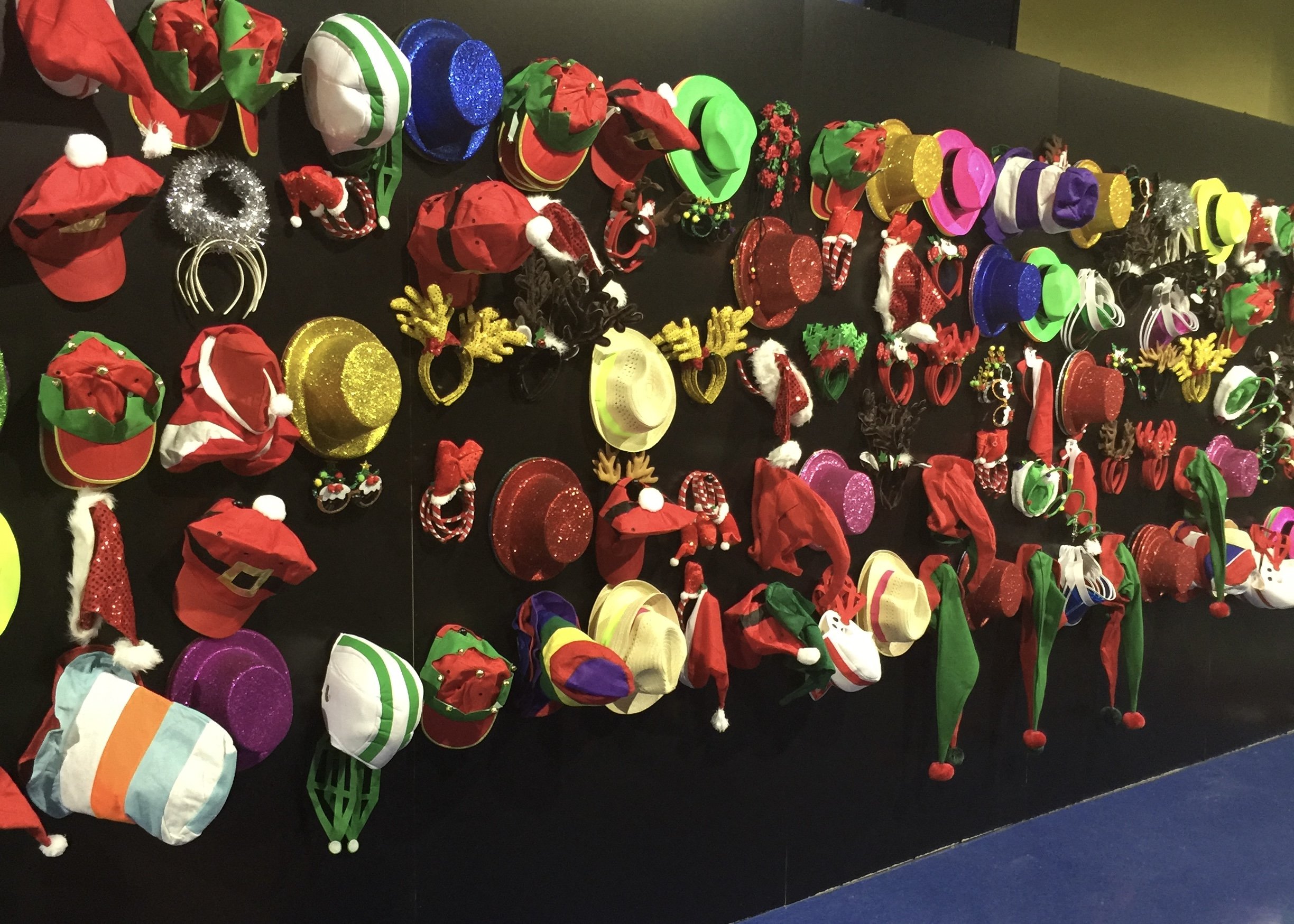 A display of various colorful Christmas hats, headbands, and accessories on a black background, including Santa hats, elf hats, reindeer antlers, and glittery hats.