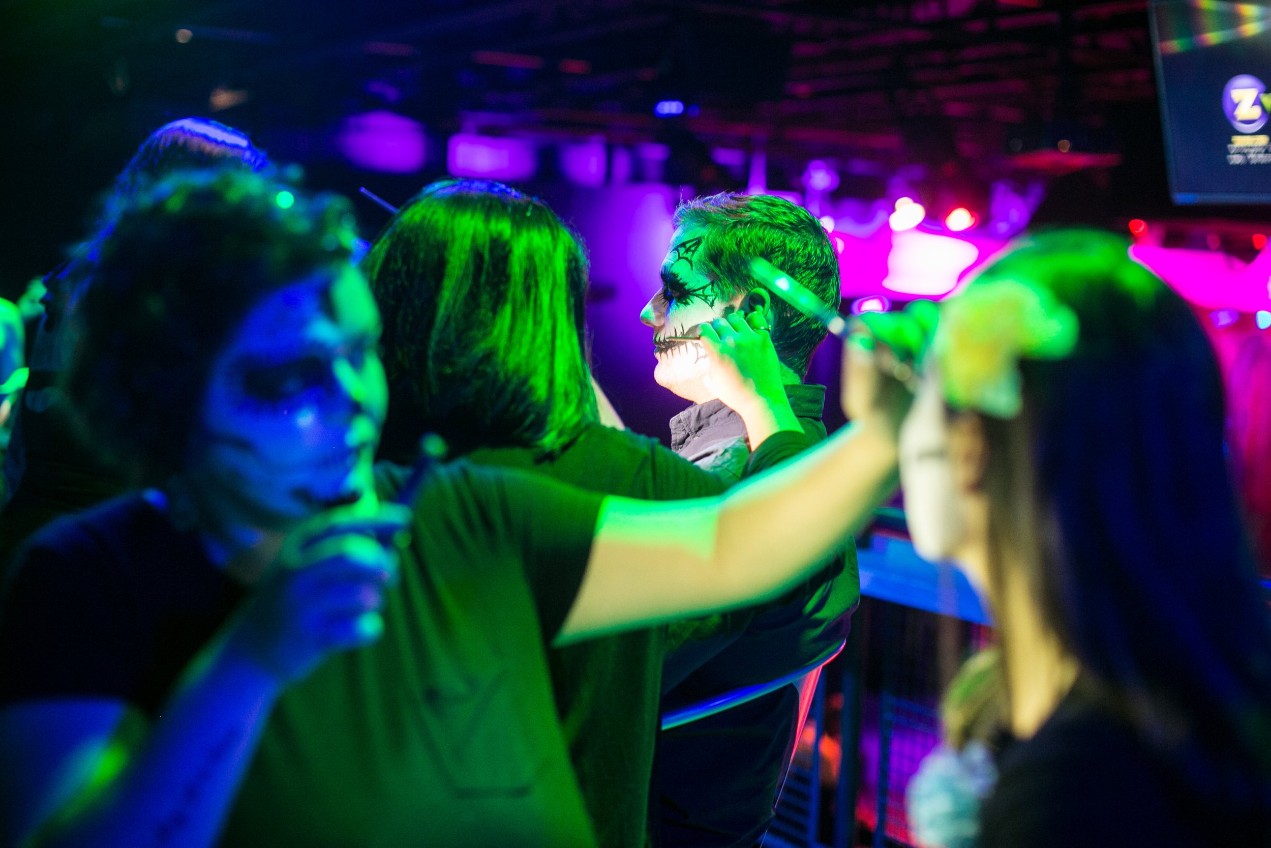 People at a party with face paint, under colorful neon lights, including someone with Halloween or Day of the Dead face makeup.