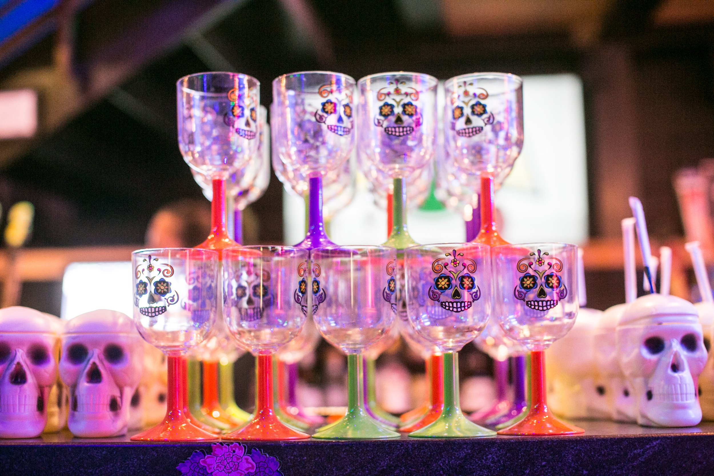 Stacked colorful glasses with sugar skull designs, each with bright, neon-colored stems, surrounded by small white skull decorations on a dark surface under vibrant lighting.