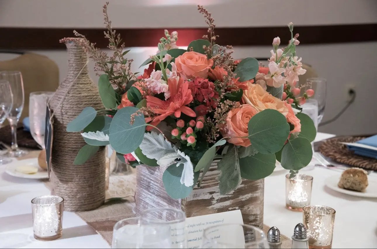 A floral centerpiece with pink roses, orange lilies, and assorted greenery on a decorated table surrounded by candles and tableware.