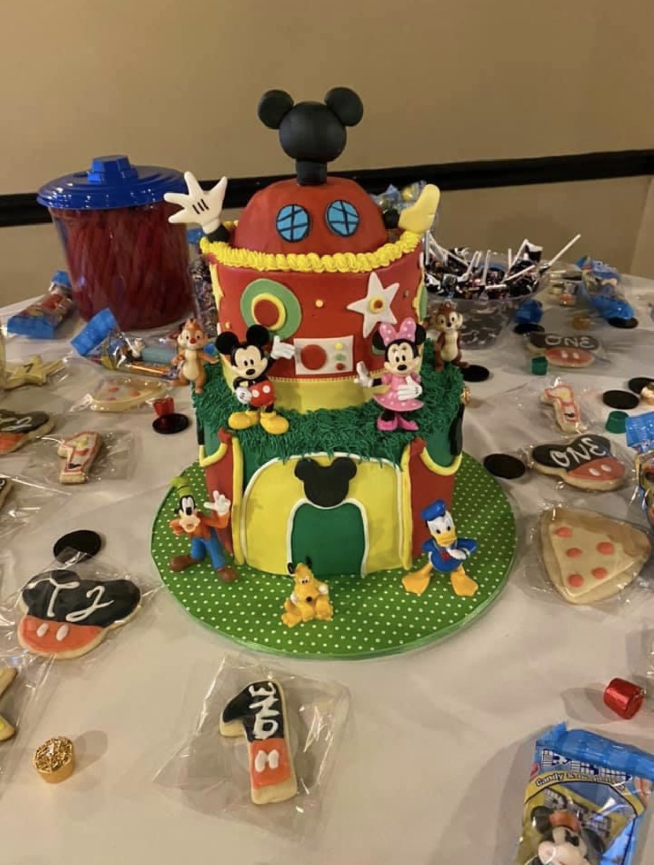 Colorful birthday cake with Disney characters and Minnie Mouse ears on the top, surrounded by candy and decorated cookies on a table.