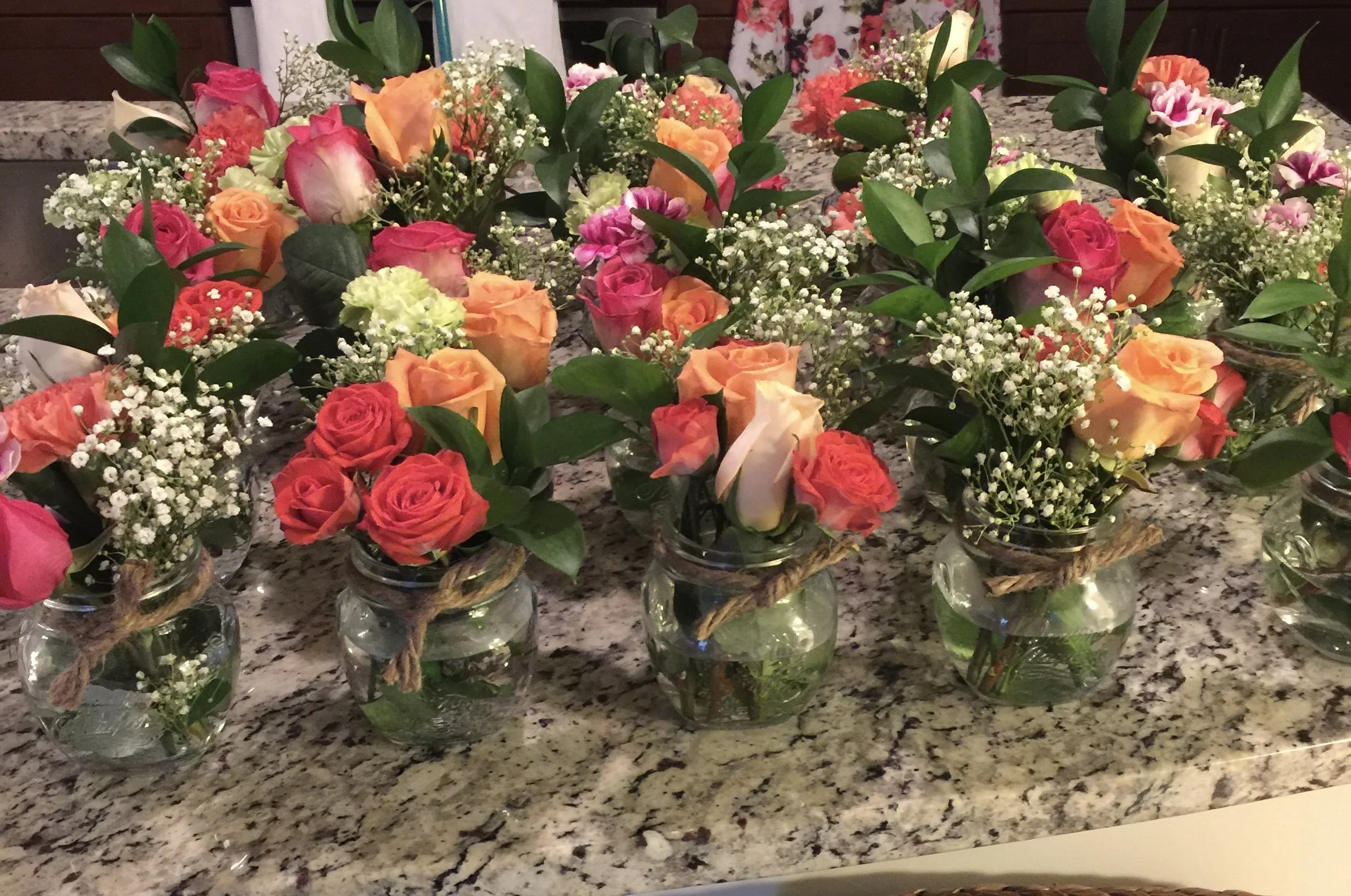 Multiple small glass jars filled with colorful roses, baby's breath, and greenery on a granite countertop.