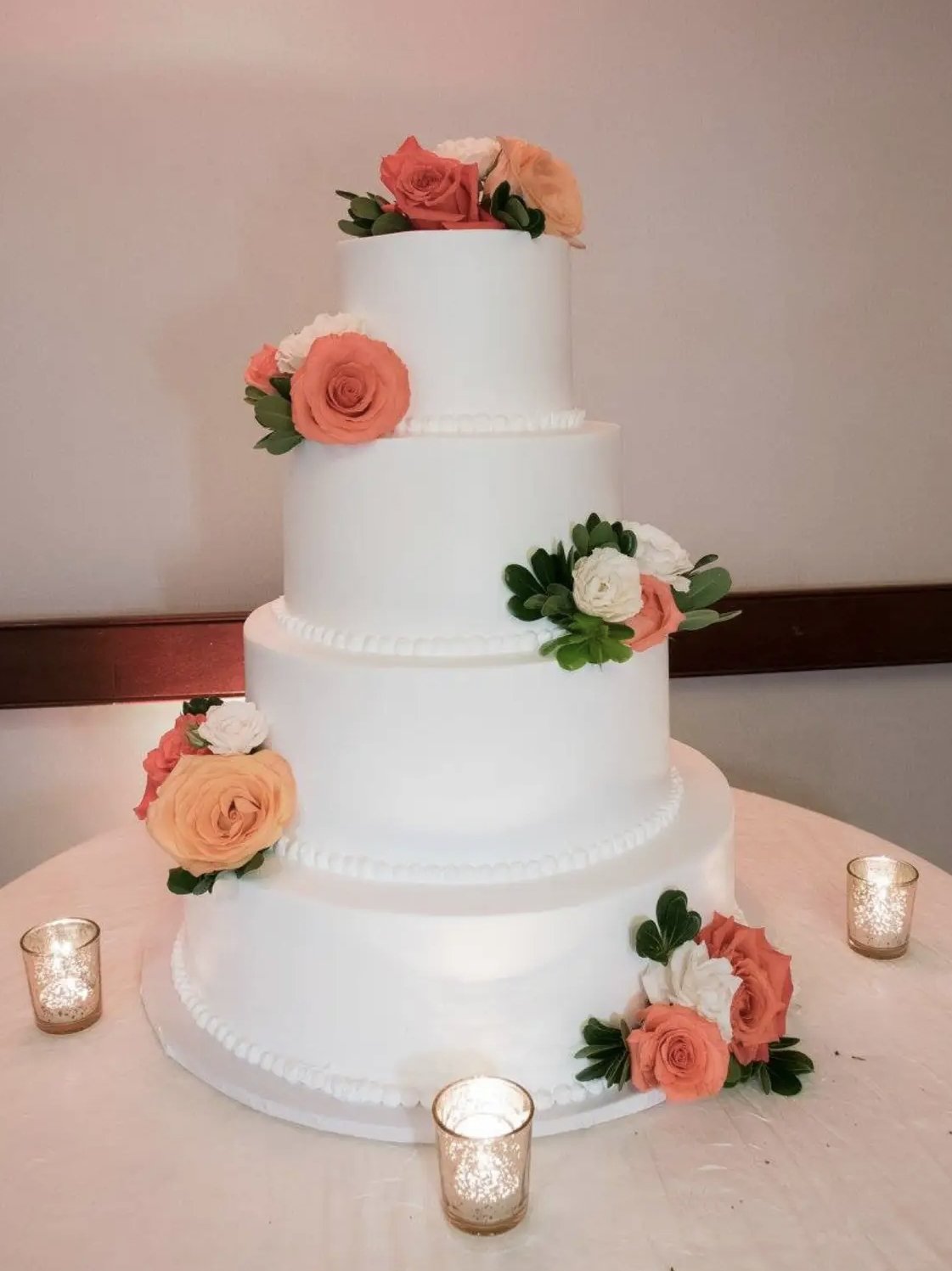 A large, four-tier white wedding cake decorated with pink, white, and peach roses, along with green foliage, and lit by four small candles.