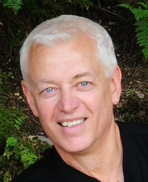 A smiling older man with short white hair, blue eyes, and earrings, outdoors with green foliage in the background.