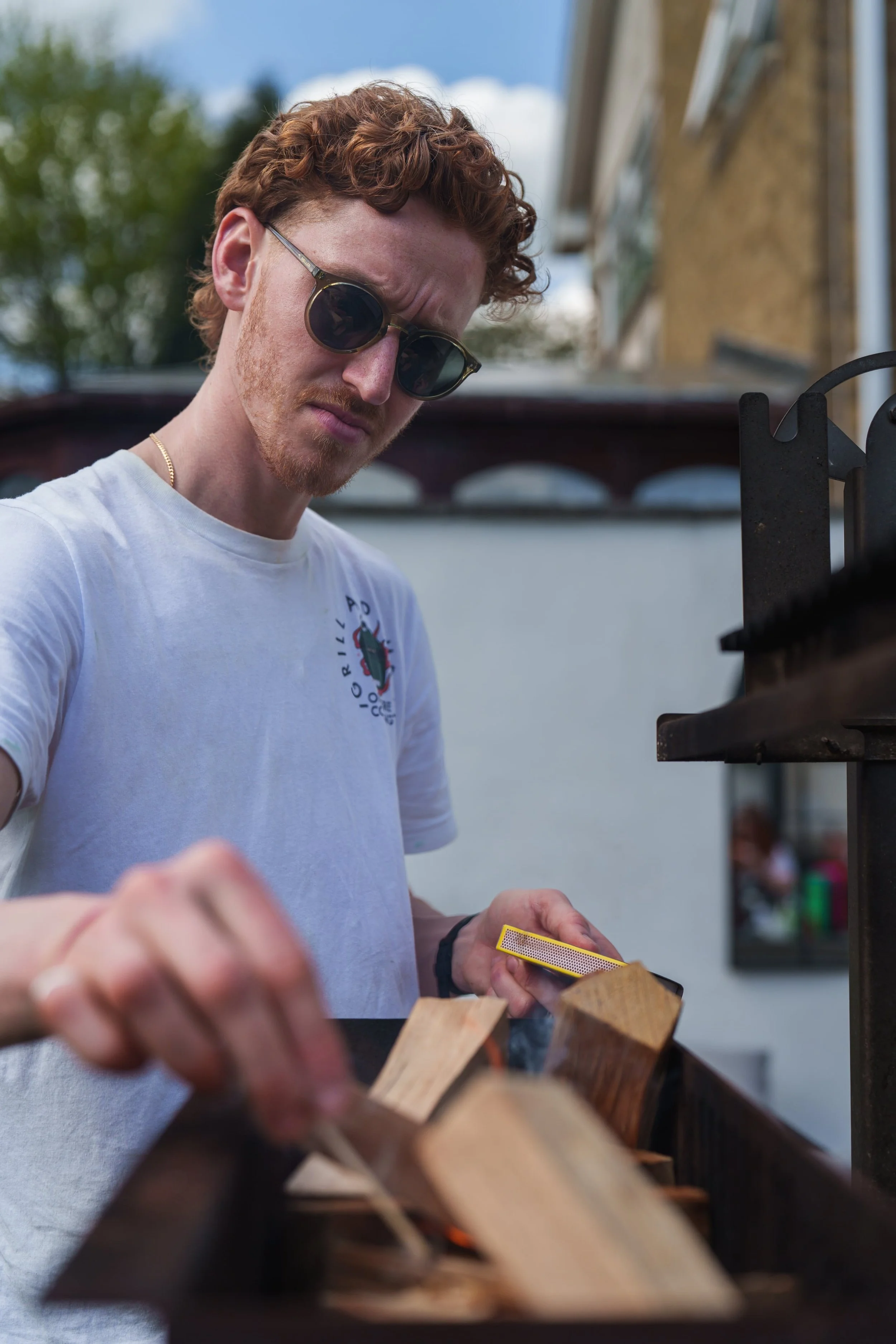 A young man with curly red hair and sunglasses lighting a barbecue grill with wooden firestarter blocks, outdoors on a bright day.