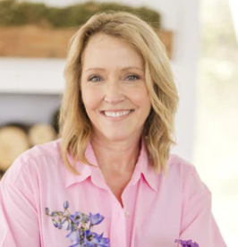 A woman with blonde hair and a pink shirt smiling in a bright, indoor setting with a blurred background.
