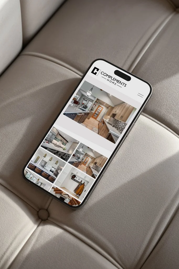 Smartphone displaying a website for home complements with images of modern kitchen and living room interior designs, placed on a beige cushioned surface.