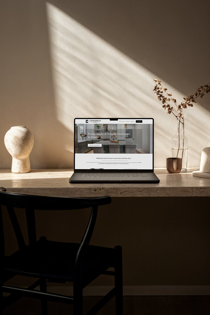 Laptop on a beige desk displaying a website for a design studio and general contractors in Oregon. The desk has decorative items including a textured vase, a small ceramic pot, and a glass vase with dried branches. Sunlight creates striped shadows on the wall.