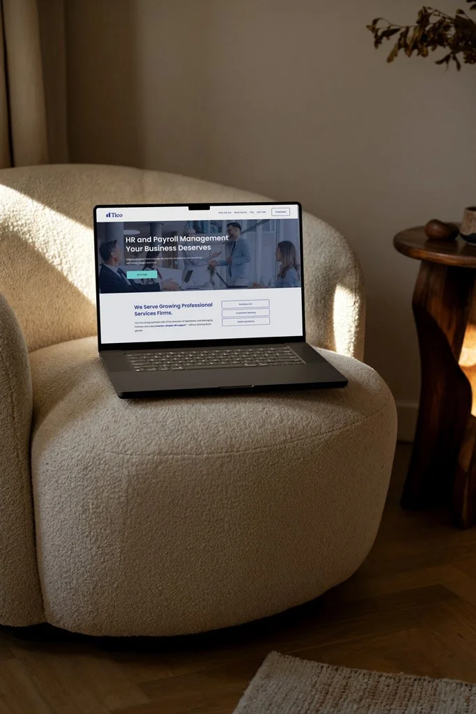Laptop on a cozy beige armchair displaying a website related to HR and payroll management, in a living room setting with a side table and a decorative plant.
