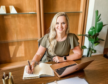 A woman sitting at a desk with an open notebook, a pen, and a laptop, smiling at the camera.