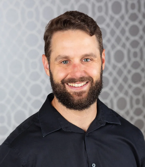 A man with short dark hair and a beard smiling, wearing a black shirt, with a grey patterned background.