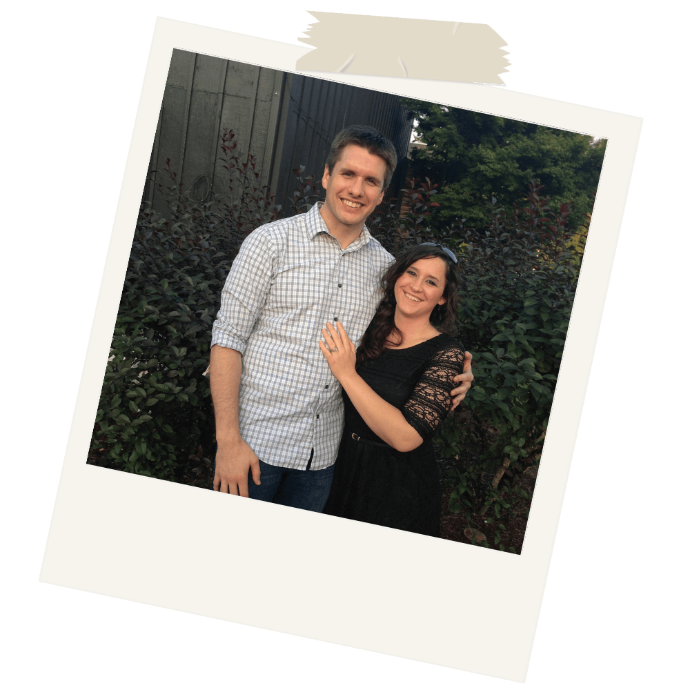 A smiling man and woman posing together outdoors in front of bushes and trees, with the woman showing her ring on her left hand.