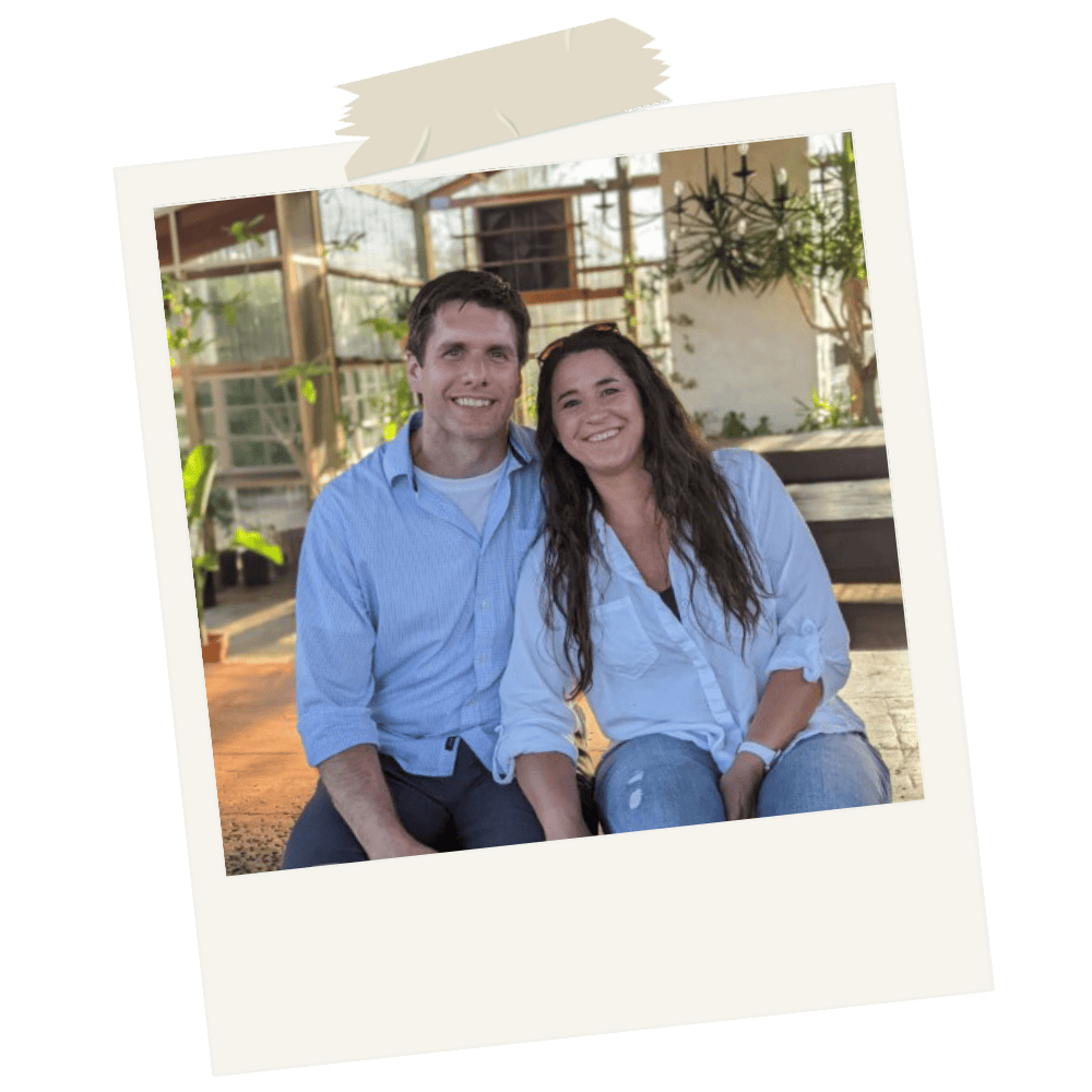 A smiling man and woman sit close together inside a greenhouse with plants and large windows, posing for a photo.