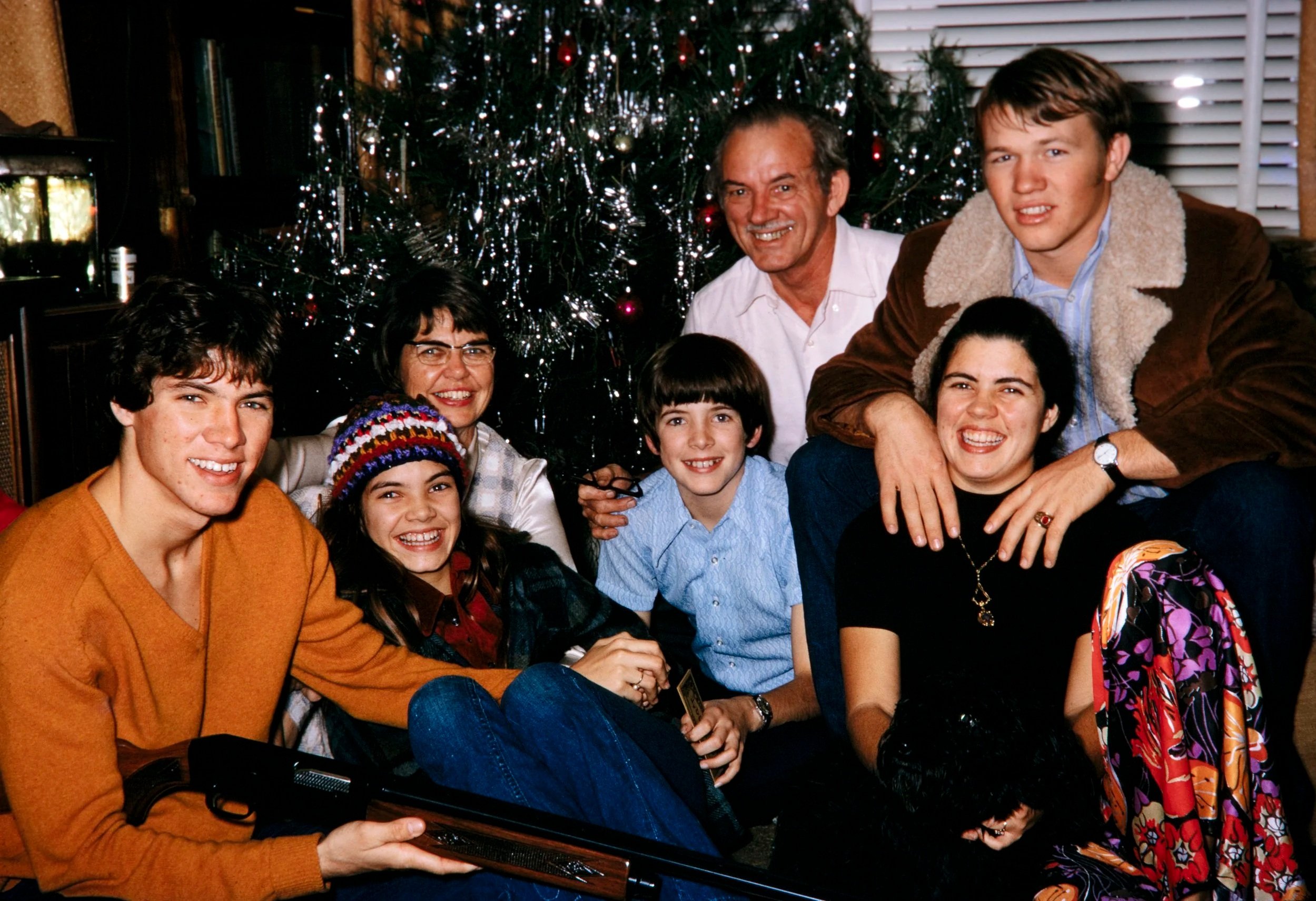 Family celebrating Christmas around a decorated tree, with people smiling and sitting together indoors.