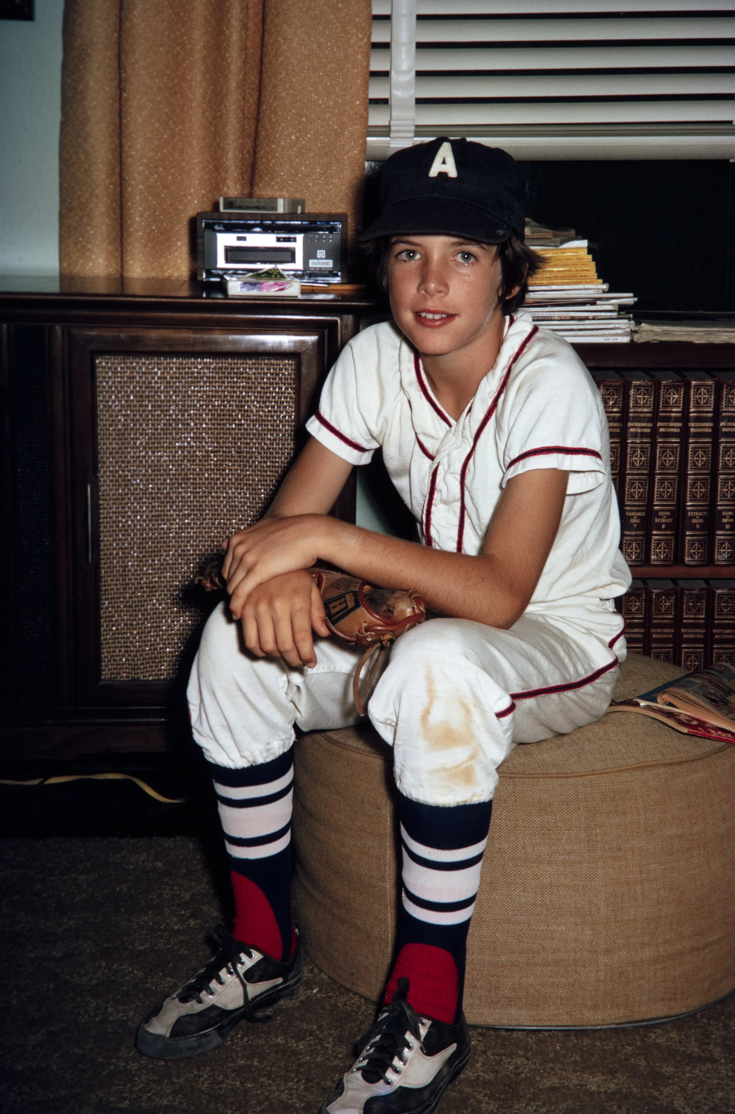 A boy sitting on a brown ottoman in a living room, wearing a baseball uniform with a navy cap, white shirt, white pants, and striped socks with sneakers. He is holding a baseball glove, with a stack of magazines on the ottoman beside him and a wooden cabinet behind him.