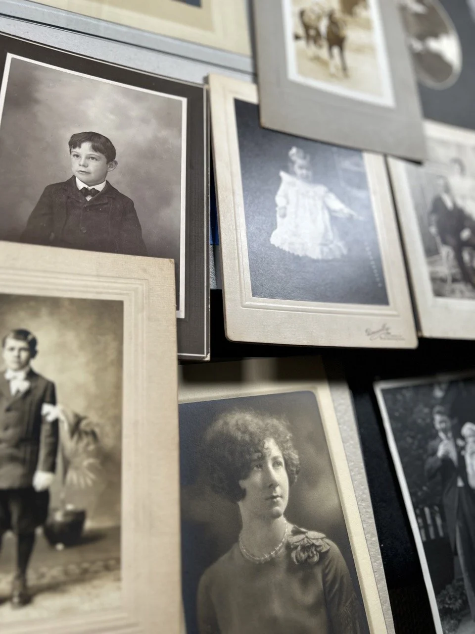 A collection of vintage black and white photographs of people, including a young boy in a suit, a woman with curly hair, and a man in historical attire, arranged on a surface.