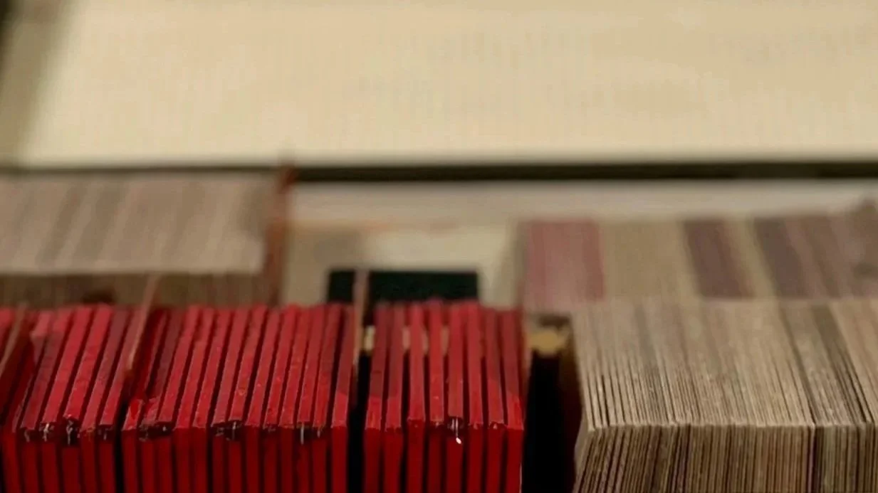 A blurry image showing a row of red books or possibly DVDs stacked vertically and some other items with brownish and beige colors, with a surface or wall above them.