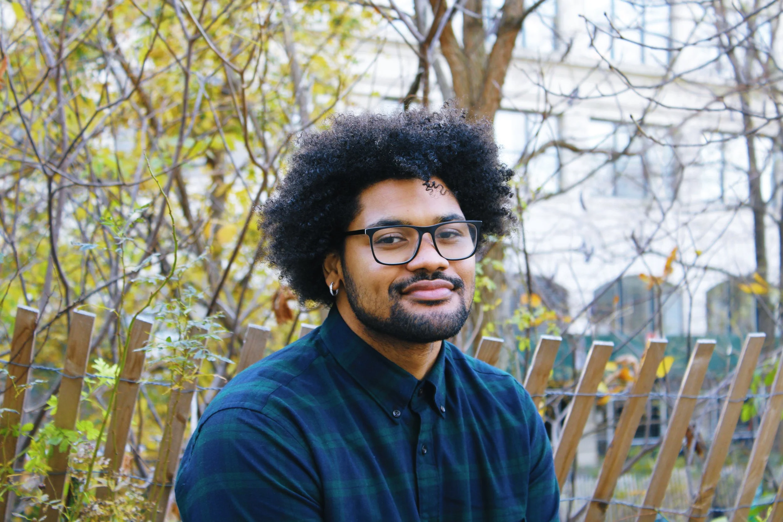A young man with curly black hair, glasses, and a beard, wearing a dark blue and green plaid shirt, standing outdoors in front of a wooden fence and trees with yellow and green leaves.