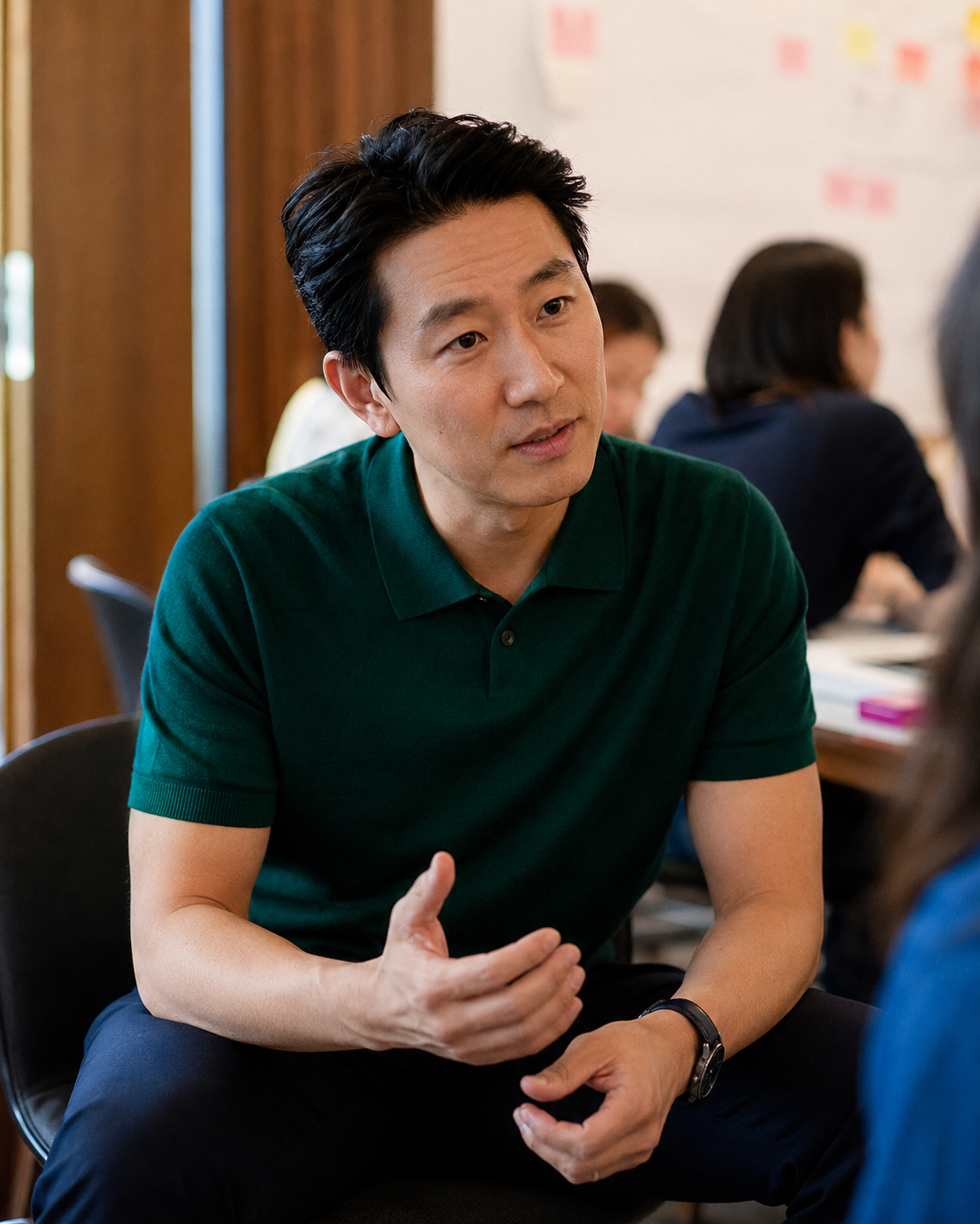 A man with black hair wearing a dark green polo shirt, engaged in conversation, gesturing with his right hand, seated in a room with other people in the background.