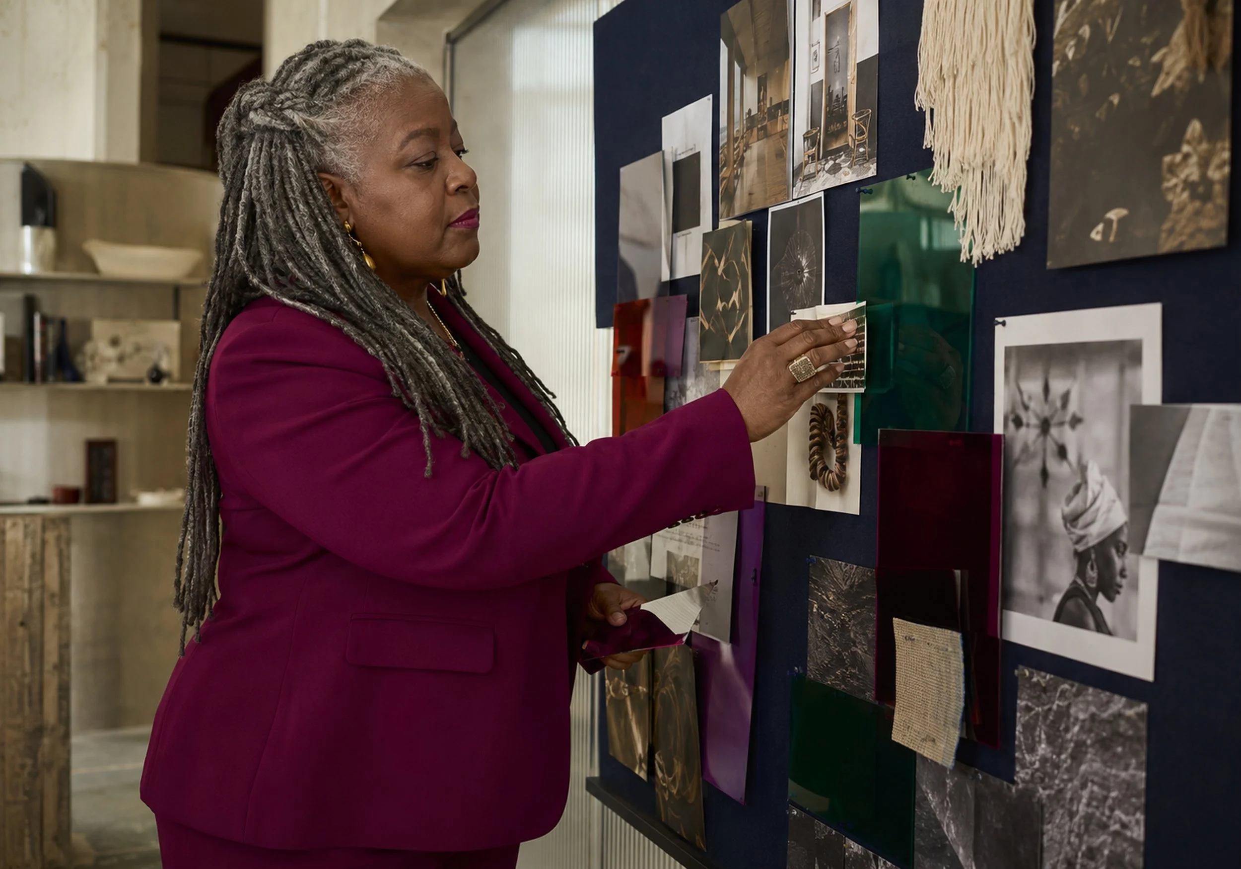 An older woman with gray dreadlocks and gold earrings wearing a magenta blazer looking at photos and fabric samples on a display board.