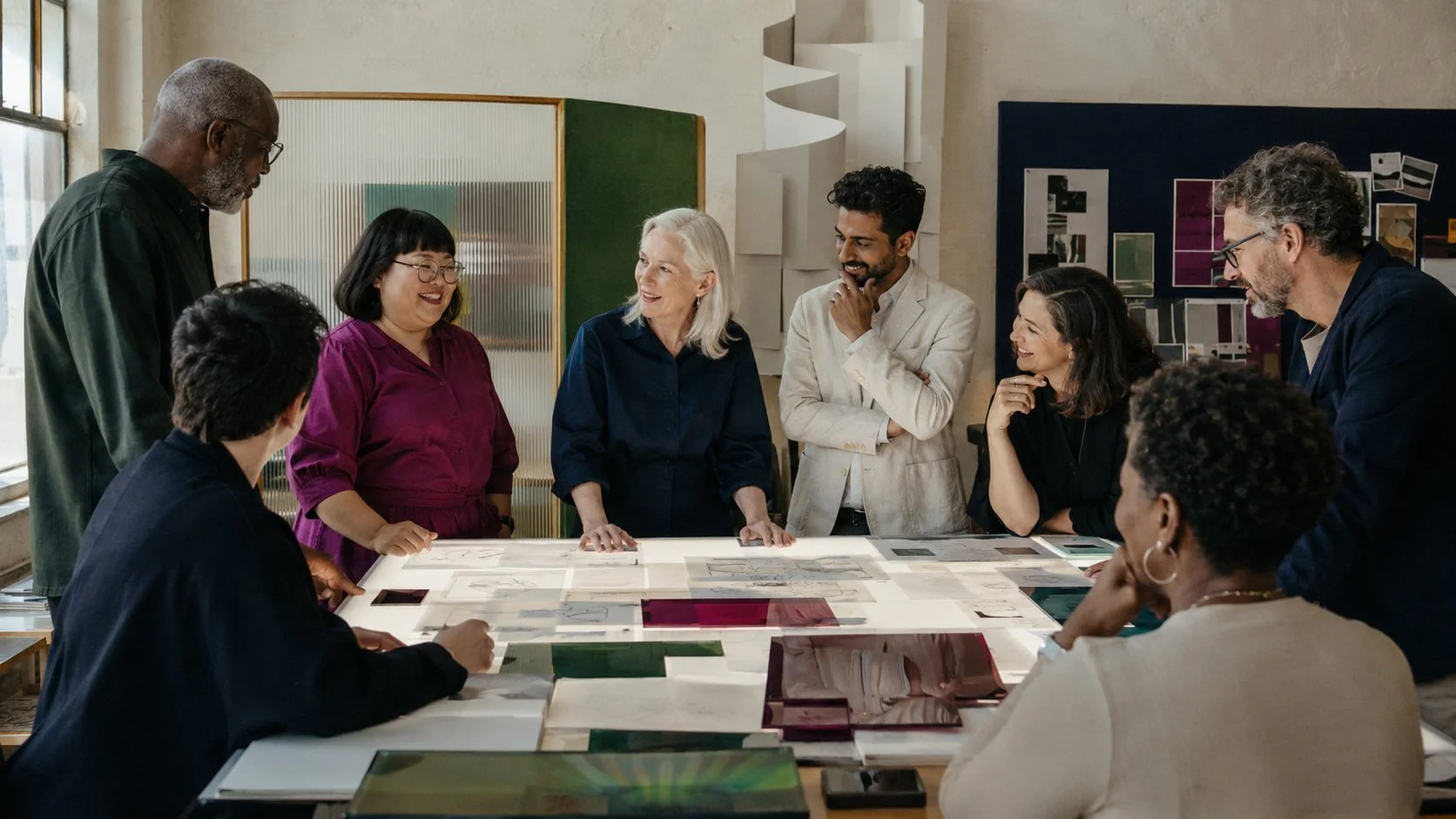A diverse group of ten people gathered around a table in a modern office, engaging in a discussion and smiling.