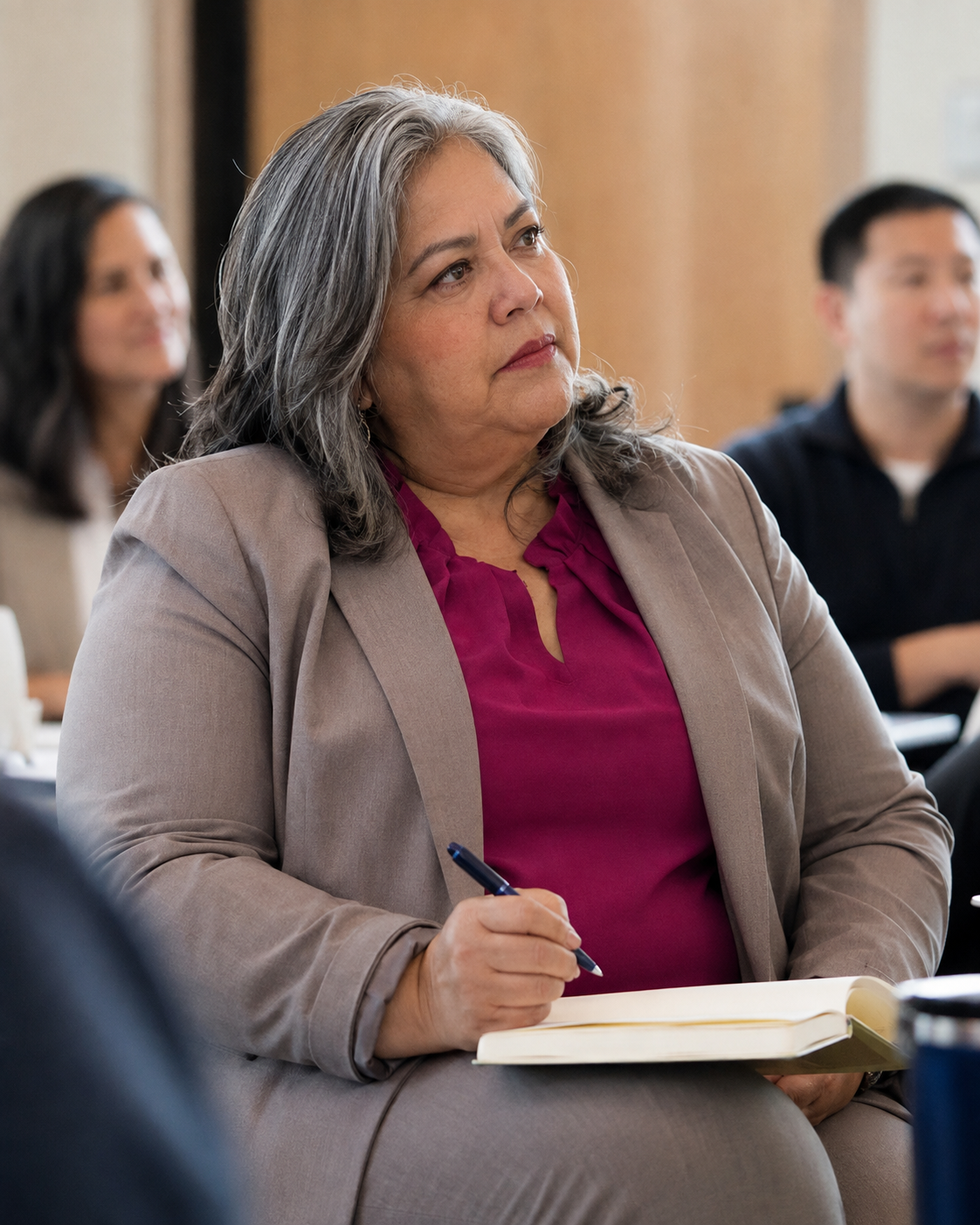 A woman with gray hair, wearing a beige blazer and magenta blouse, taking notes with a pen during a meeting or conference.