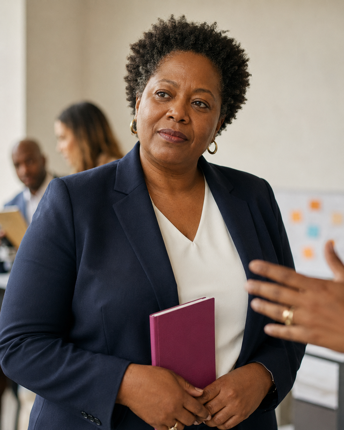 A professional woman with short curly hair, wearing a navy blazer and white shirt, holding a purple notebook, engaged in conversation at a meeting or conference.