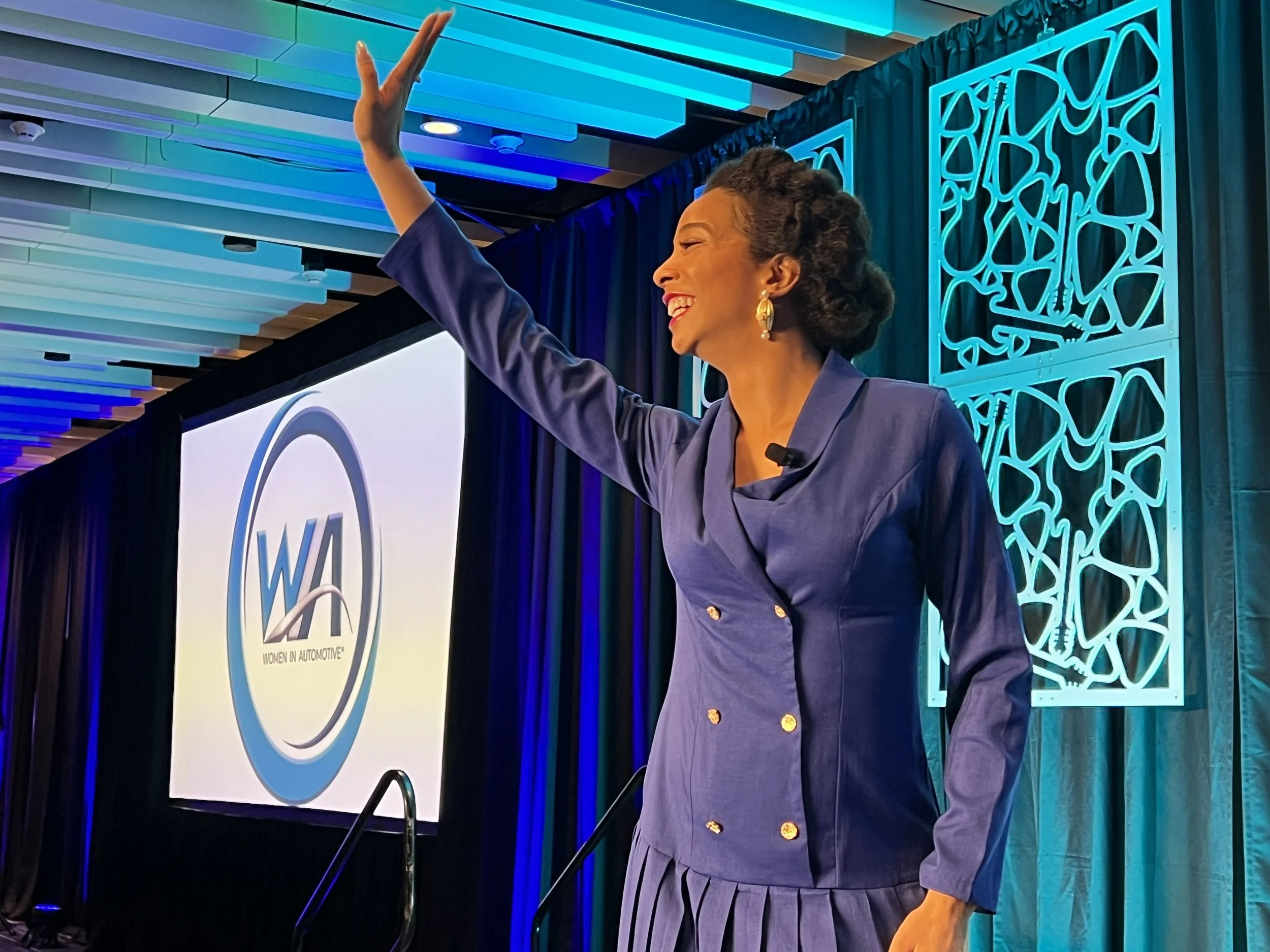 A woman in a blue suit smiling and waving at a conference or event with a Women in Automotive sign in the background.