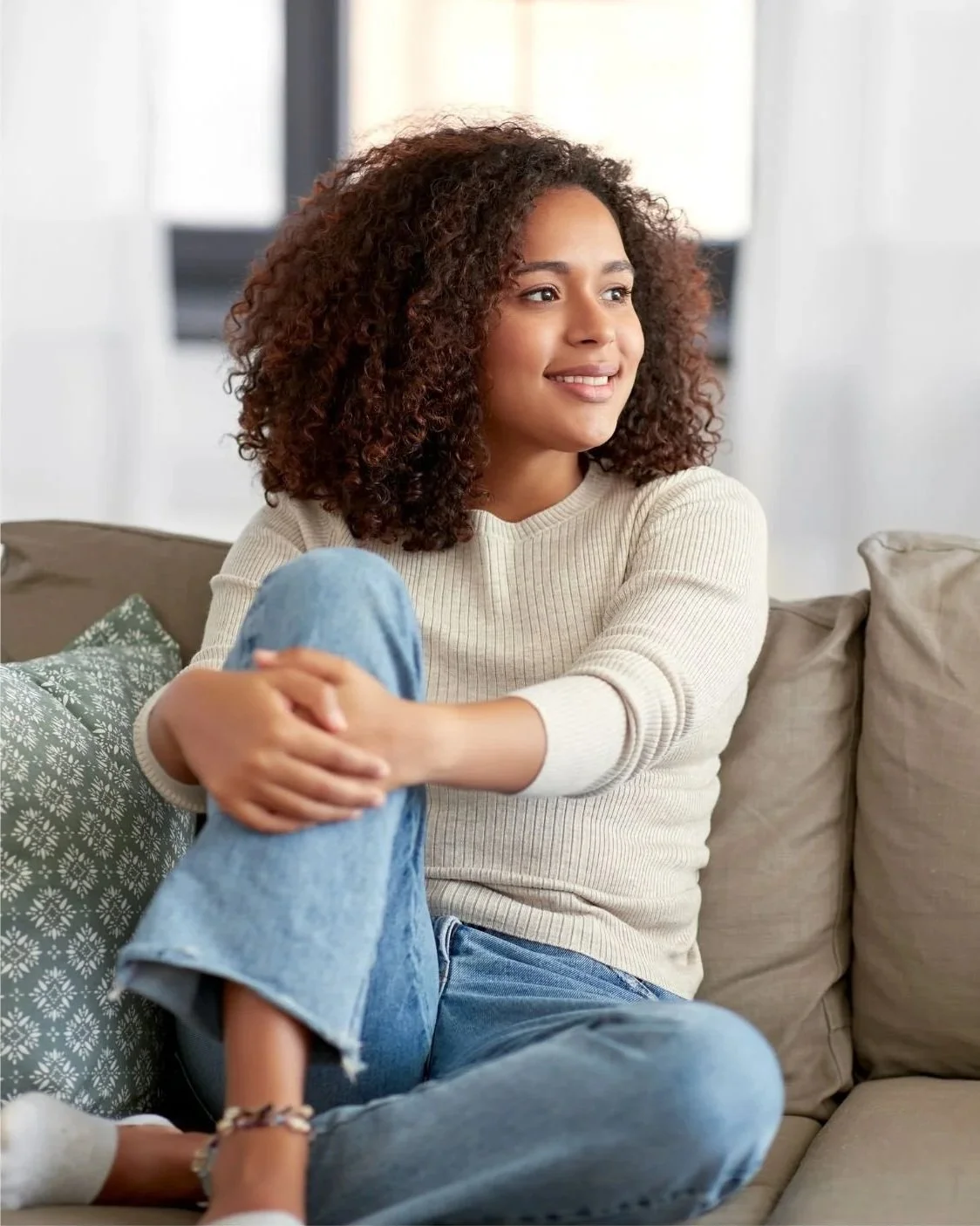 A woman with curly hair sitting on a beige sofa, smiling, wearing a cream sweater and jeans, with a patterned pillow beside her.
