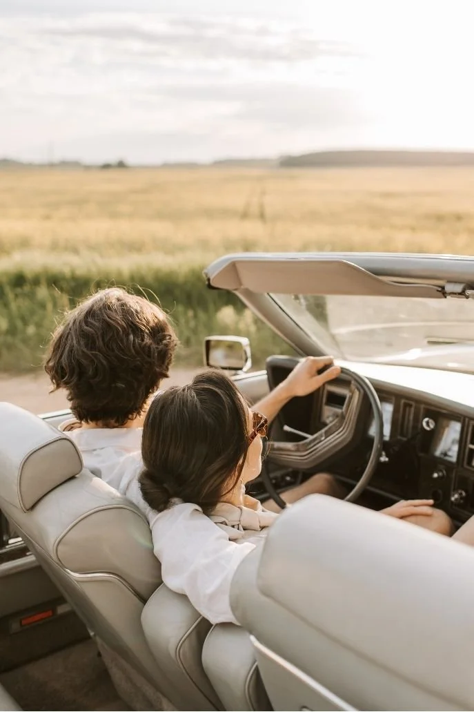Two people sitting in a classic car driving through a rural landscape with open fields and cloudy sky.