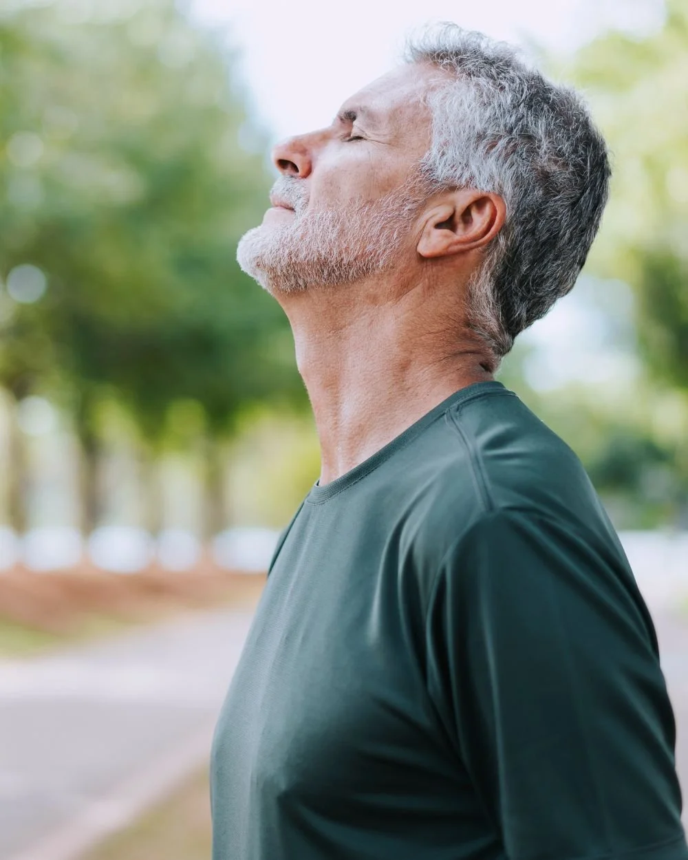 A senior man with gray hair and a beard standing outdoors with eyes closed, enjoying the fresh air on a sunny day.